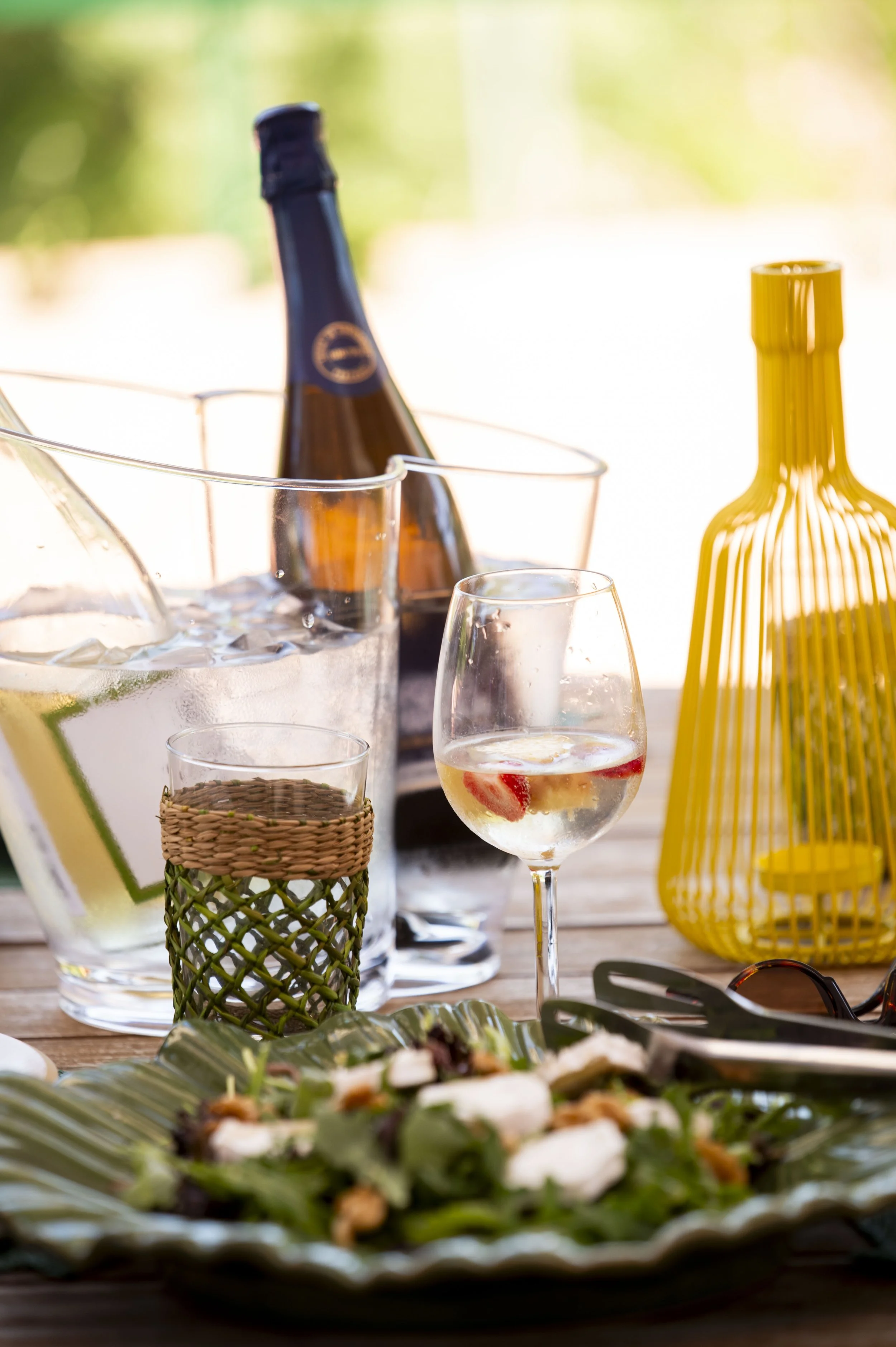 Mesa com bebidas, incluindo garrafa de cerveja, copo de drink com fruta, jarra de água com limão, e objetos decorativos amarelos.