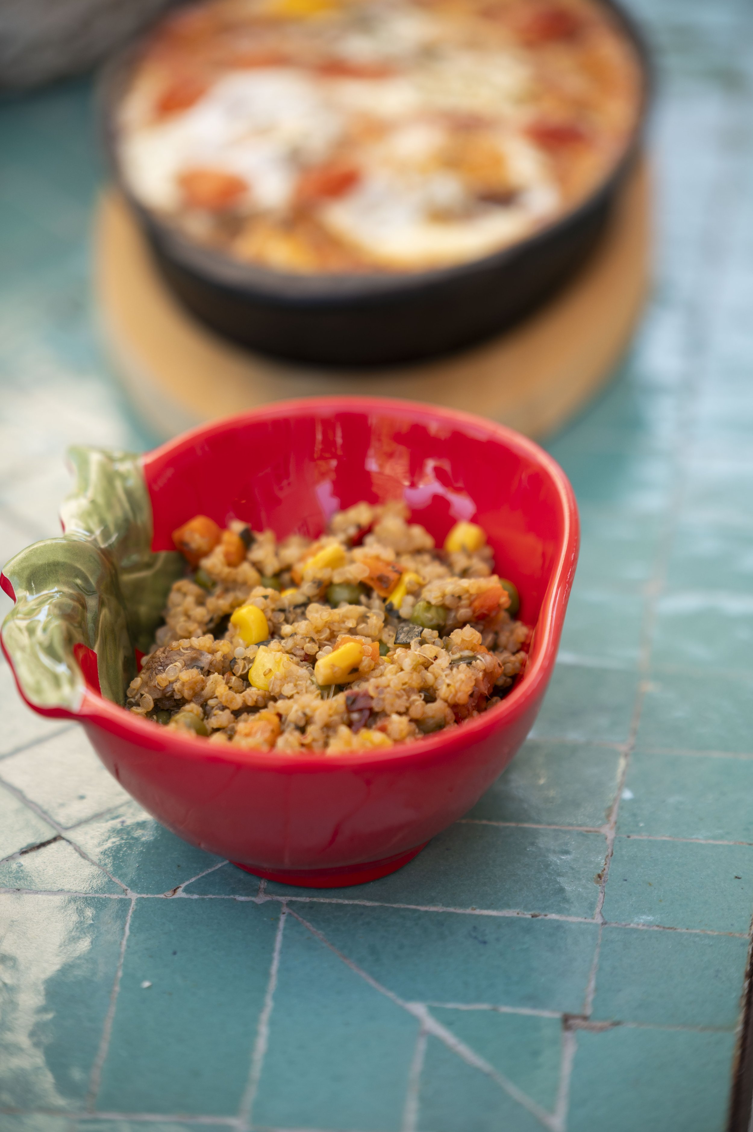Prato de arroz com legumes e carne, e uma tigela vermelha com comida no primeiro plano, em uma mesa de cerâmica azul.