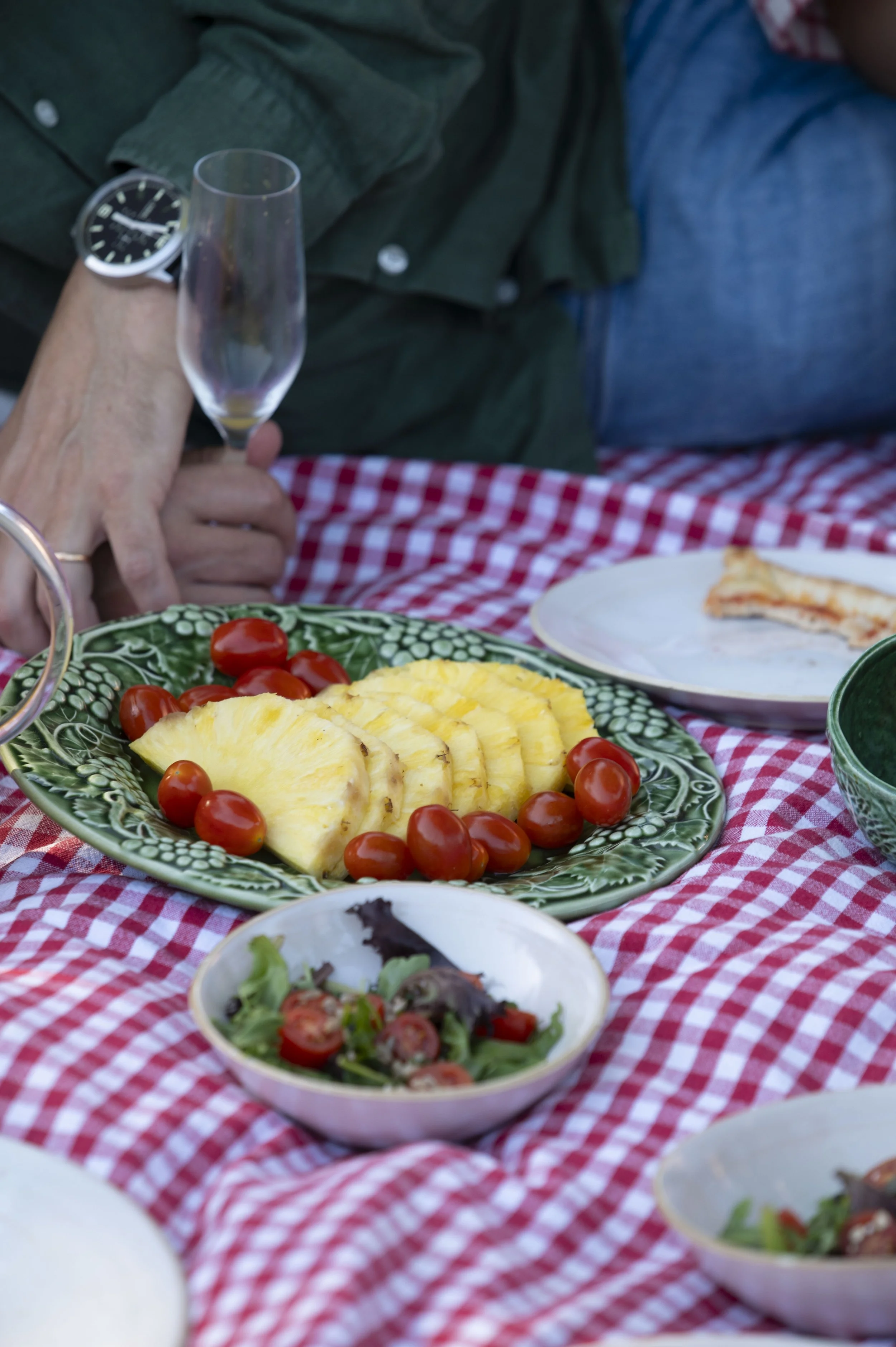 Prato com fatias de abacaxi e tomates cereja ao lado, em uma mesa com toalha de mesa xadrez vermelha e branca, com outras porções de comida ao fundo.