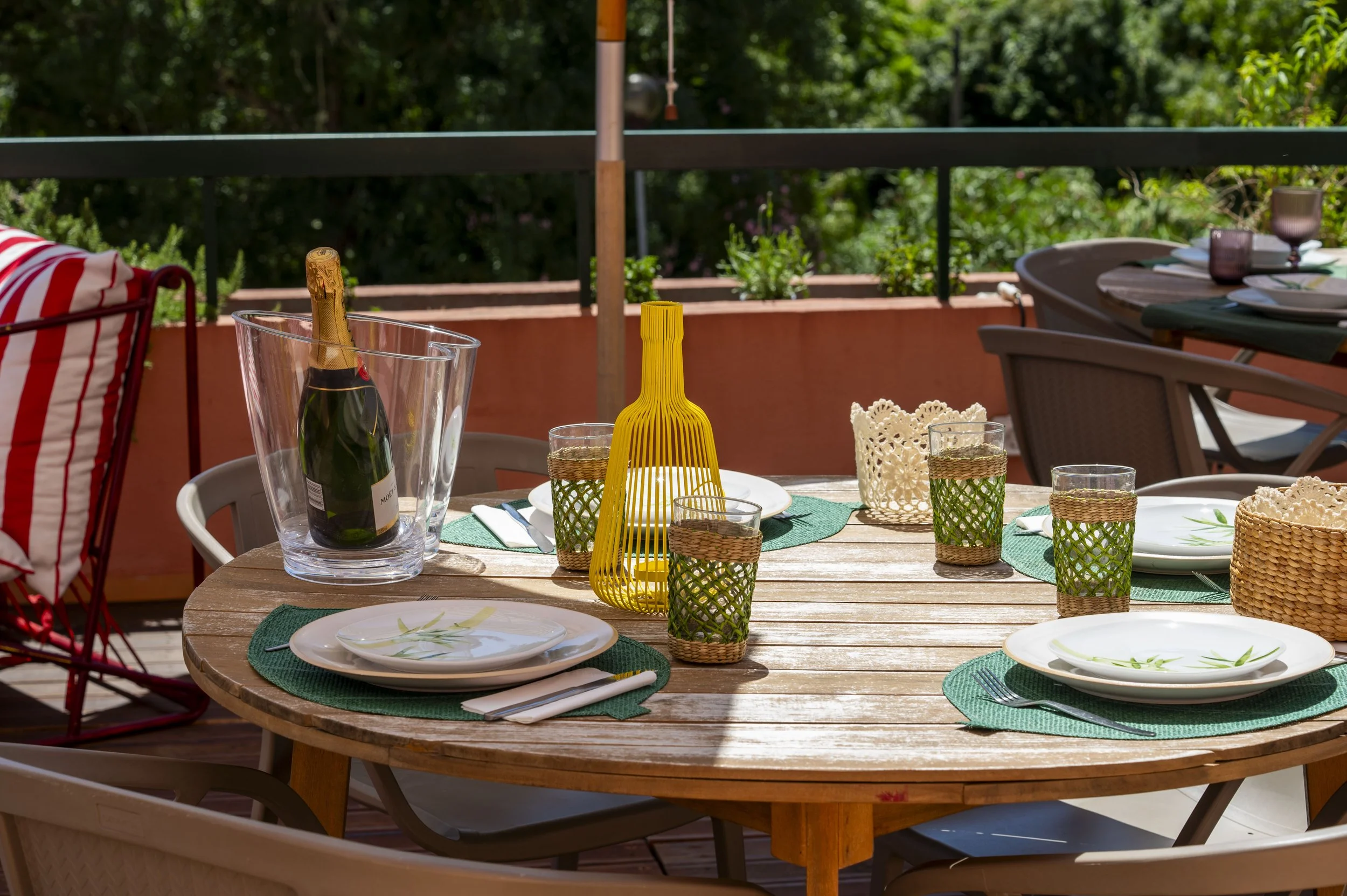 Mesa de exterior decorada com pratos, copos, uma garrafa de espumante em balde de gelo, luminária amarela, guardanapos brancos, toalhas verdes e cadeiras ao redor, com fundo de árvores verdes.