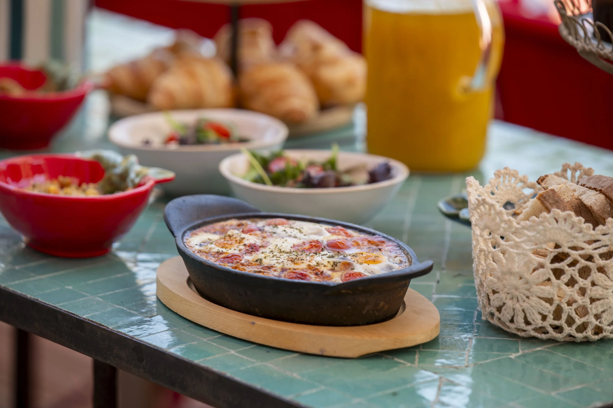 Prato de pizza com ovos, queijo e ervas, acompanhado de saladas, em uma mesa com outros pratos e pães ao fundo.