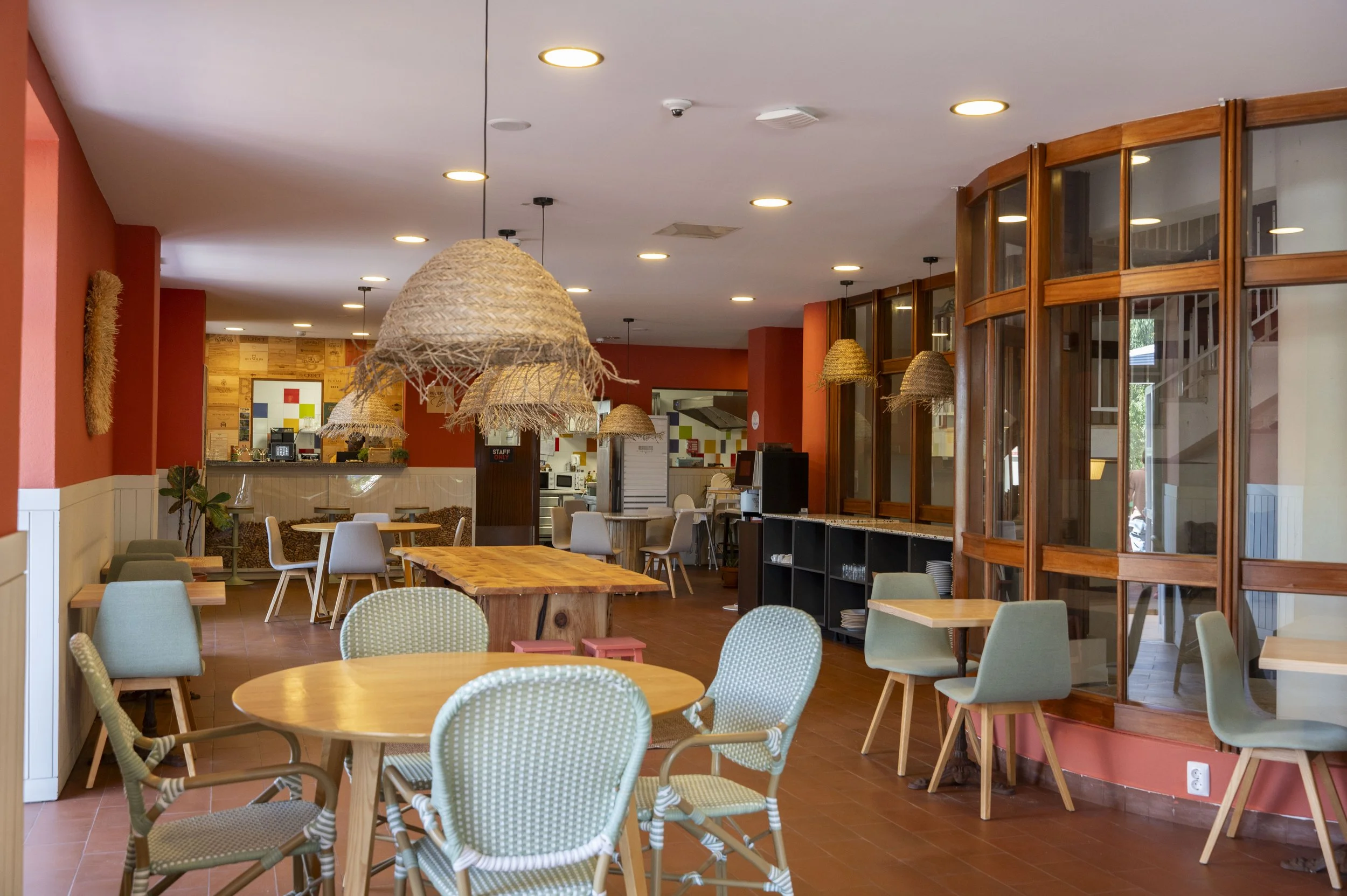 Interior de um restaurante com mesas e cadeiras modernas, luzes de teto circulares, paredes em tons de vermelho e mobiliário de madeira, decorado de forma acolhedora.