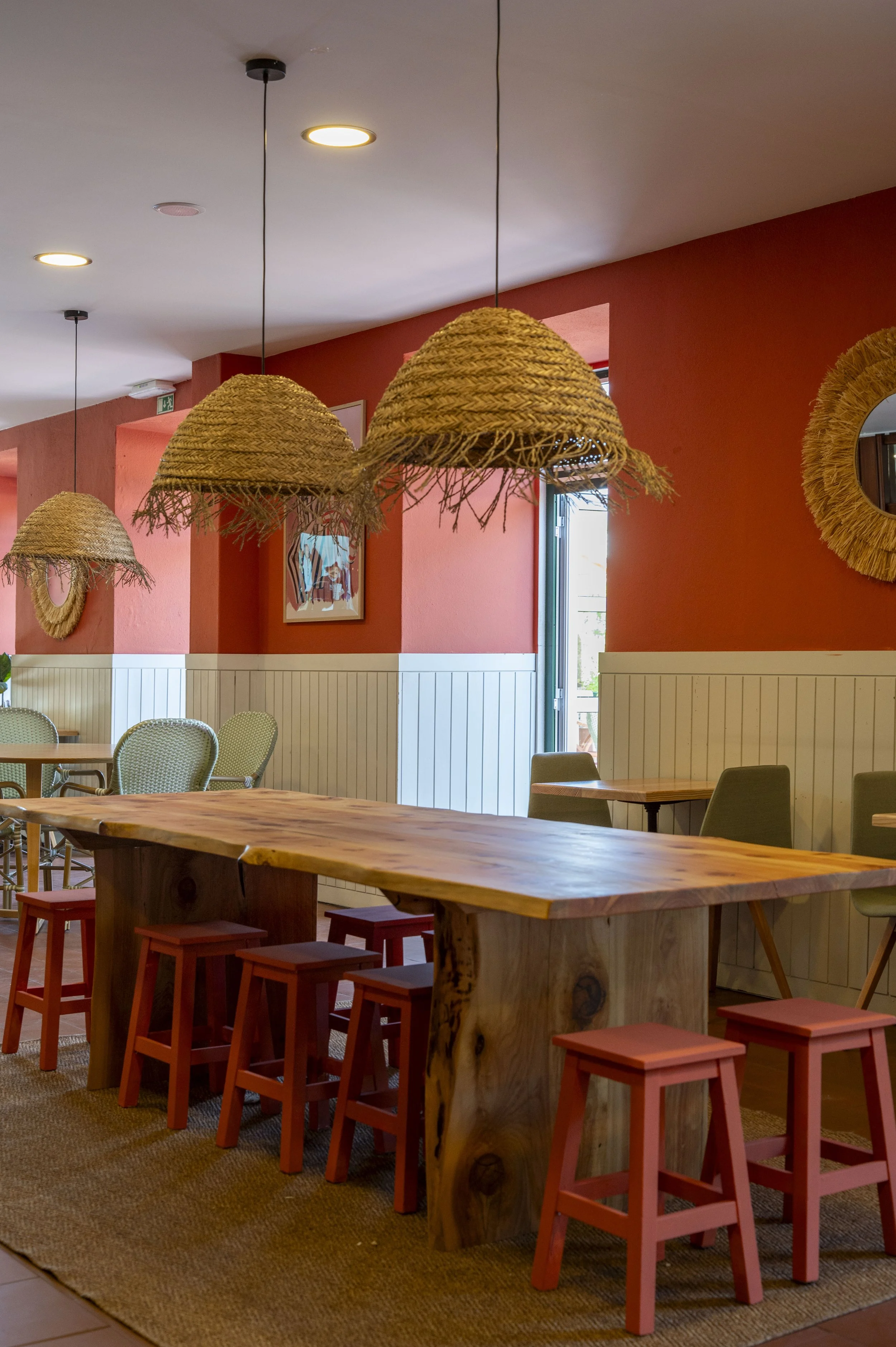 Interior de restaurante com mesa de madeira, cadeiras e banquetas vermelhas, luminárias de fibra natural penduradas no teto, parede pintada de vermelho com espelho redondo, janela ao fundo permitindo entrada de luz natural.