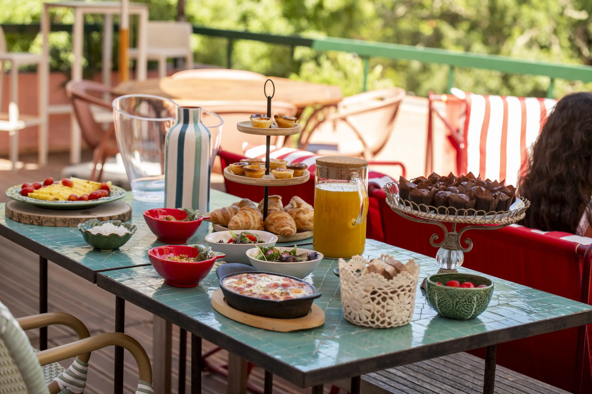 Mesa de café da manhã ao ar livre com frutas, croissants, pizza, suco, doces e utensílios, em um ambiente ensolarado com árvores ao fundo.