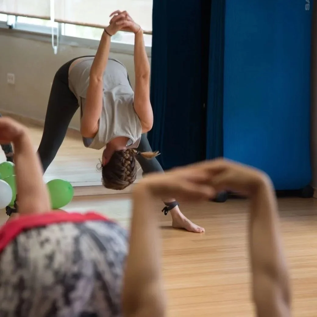 Une femme pratique le yoga en position de pont dans une salle de sport, avec des ballons verts à côté, et un enfant qui joue ou fait des activités au premier plan.