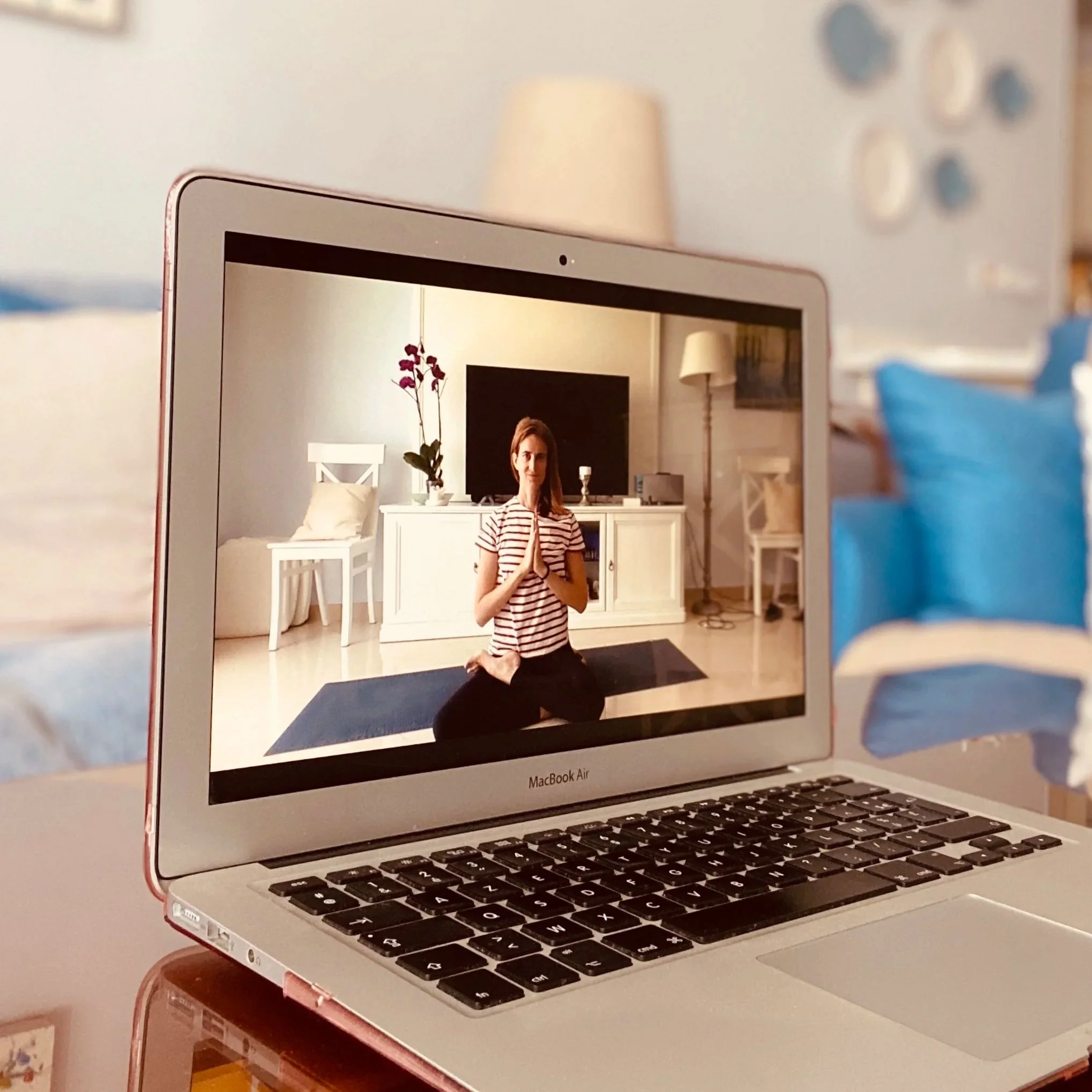 Capture d'écran d'un ordinateur portable MacBook Air avec un clavier noir, affichant une image dans l'écran d'une femme en position de prière en méditation dans un intérieur décoré avec une lampe, une table blanche, une orchidée et un mur blanc