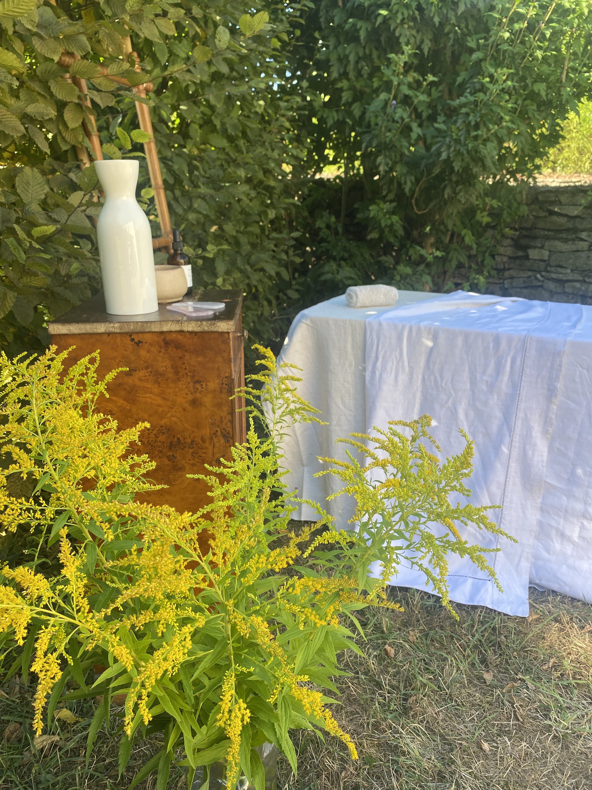Une table de massage en plein air recouverte d'une draperie blanche, avec une serviette roulée dessus et entourée de végétation verte et de fleurs jaunes, sous une lumière naturelle.