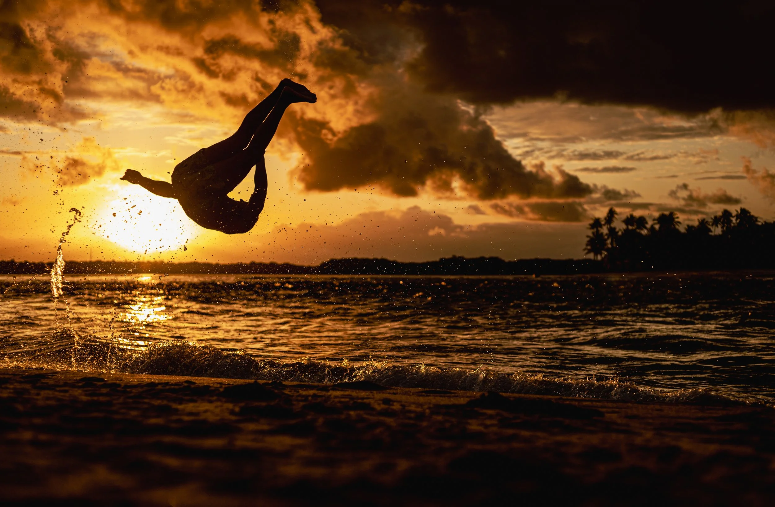 A person mid-air doing a flip over the water during sunset, with orange and dark clouds in the sky and palm trees in the distance.