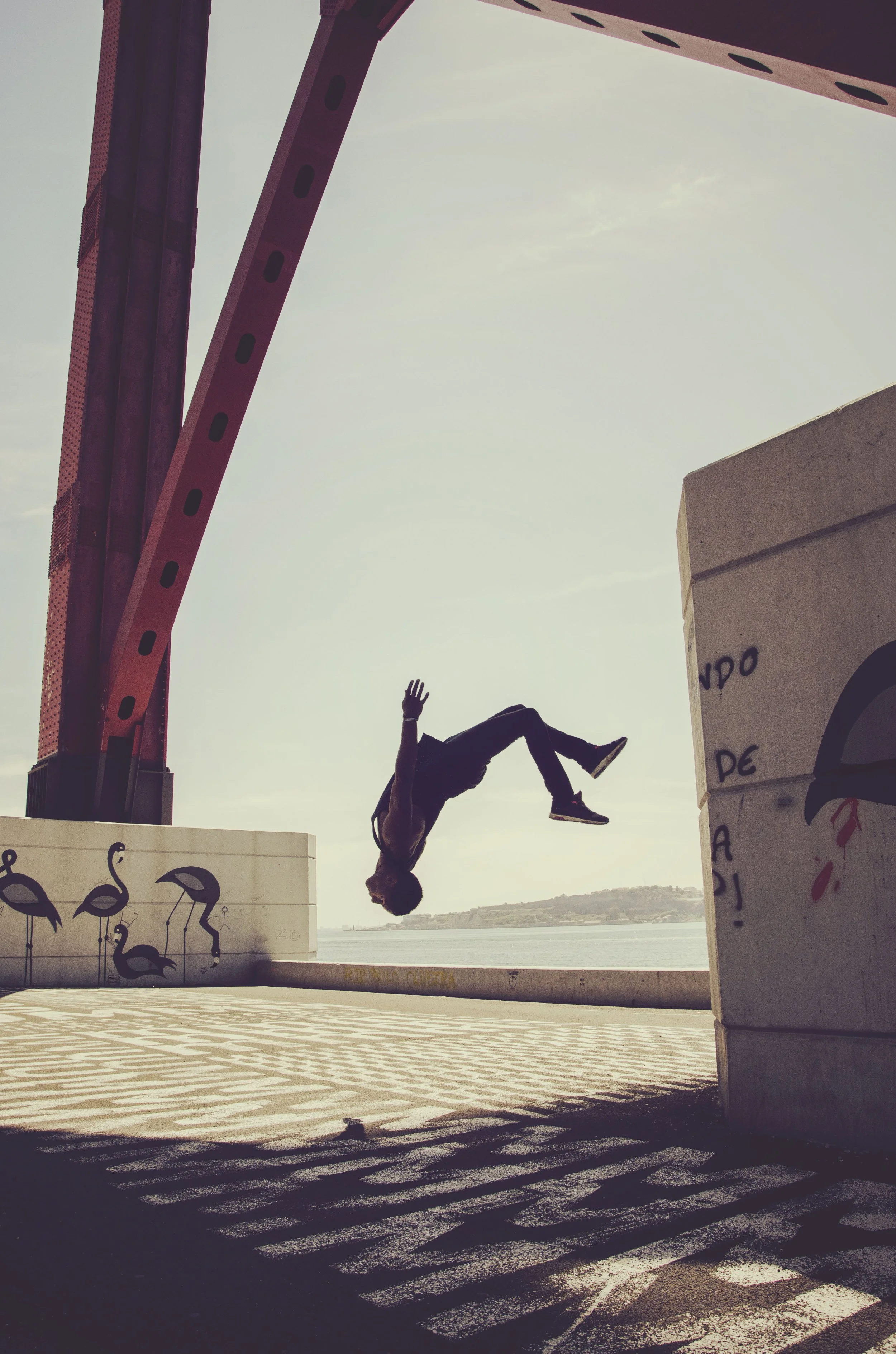 A person is captured mid-air doing a flip, with their body facing downward and legs bent, near the edge of a waterfront promenade. The silhouette is backlit by the bright sky, creating a contrast with the shadows cast on the ground.