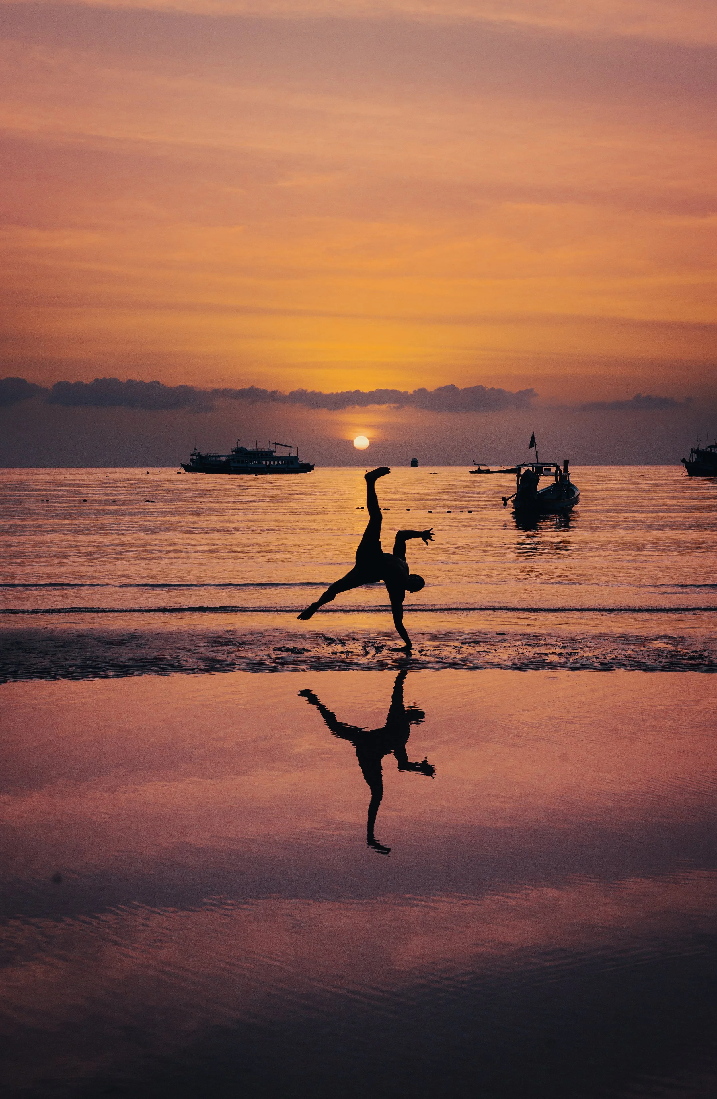 A person performing a handstand on the beach at sunset, with their reflection visible in the wet sand. Acrobatic show