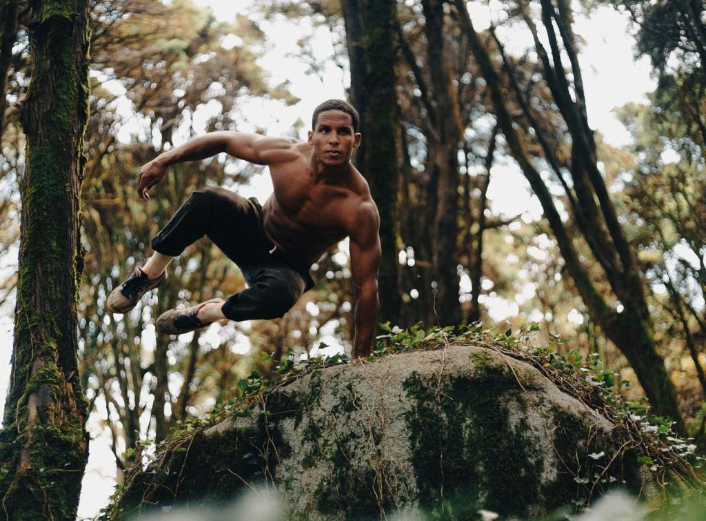 A shirtless man in athletic attire balancing on a large moss-covered rock in a forest, with trees and foliage in the background.