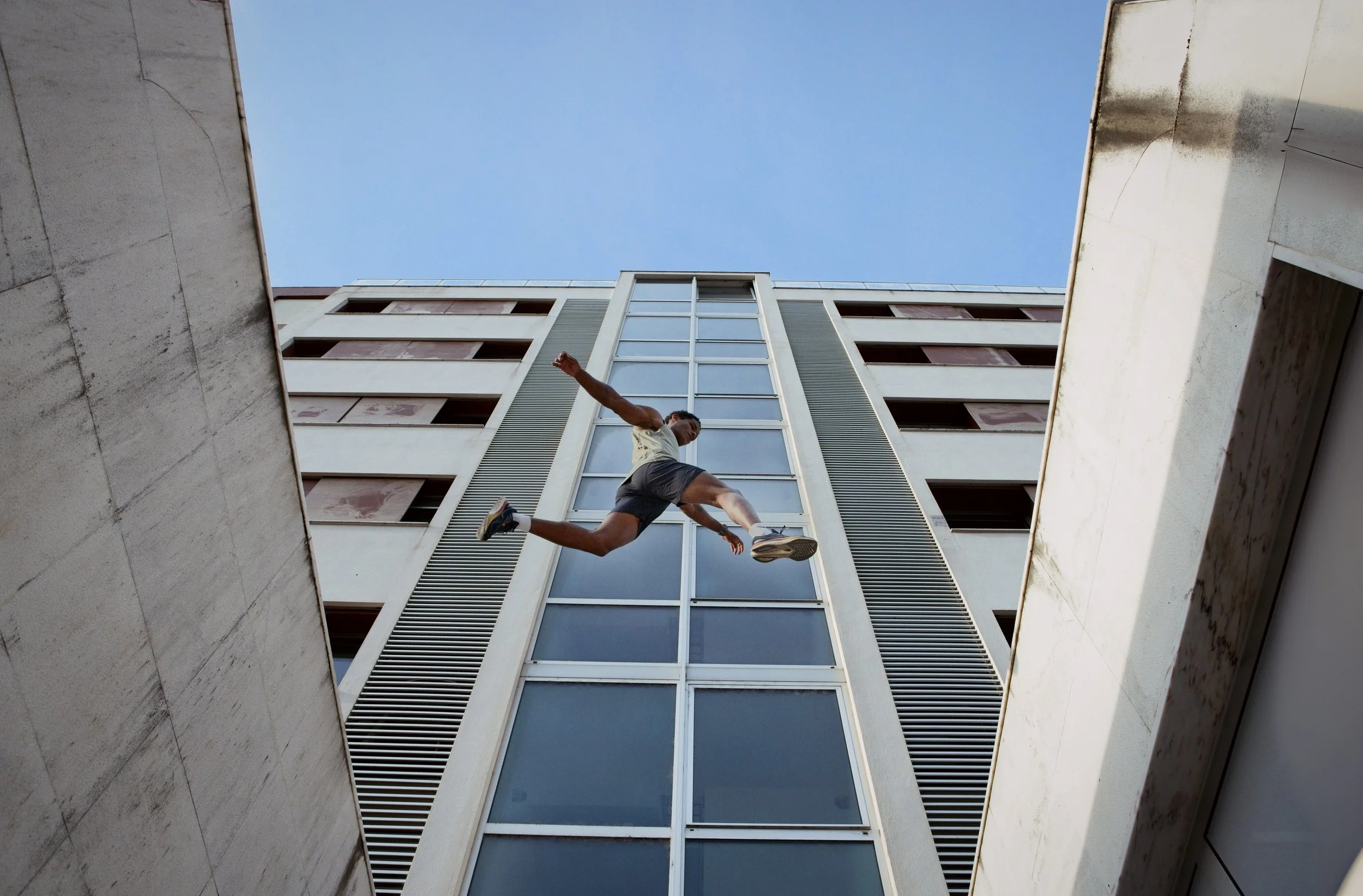 A person mid-air falling between two buildings with modern architecture and glass windows, with a blue sky in the background.