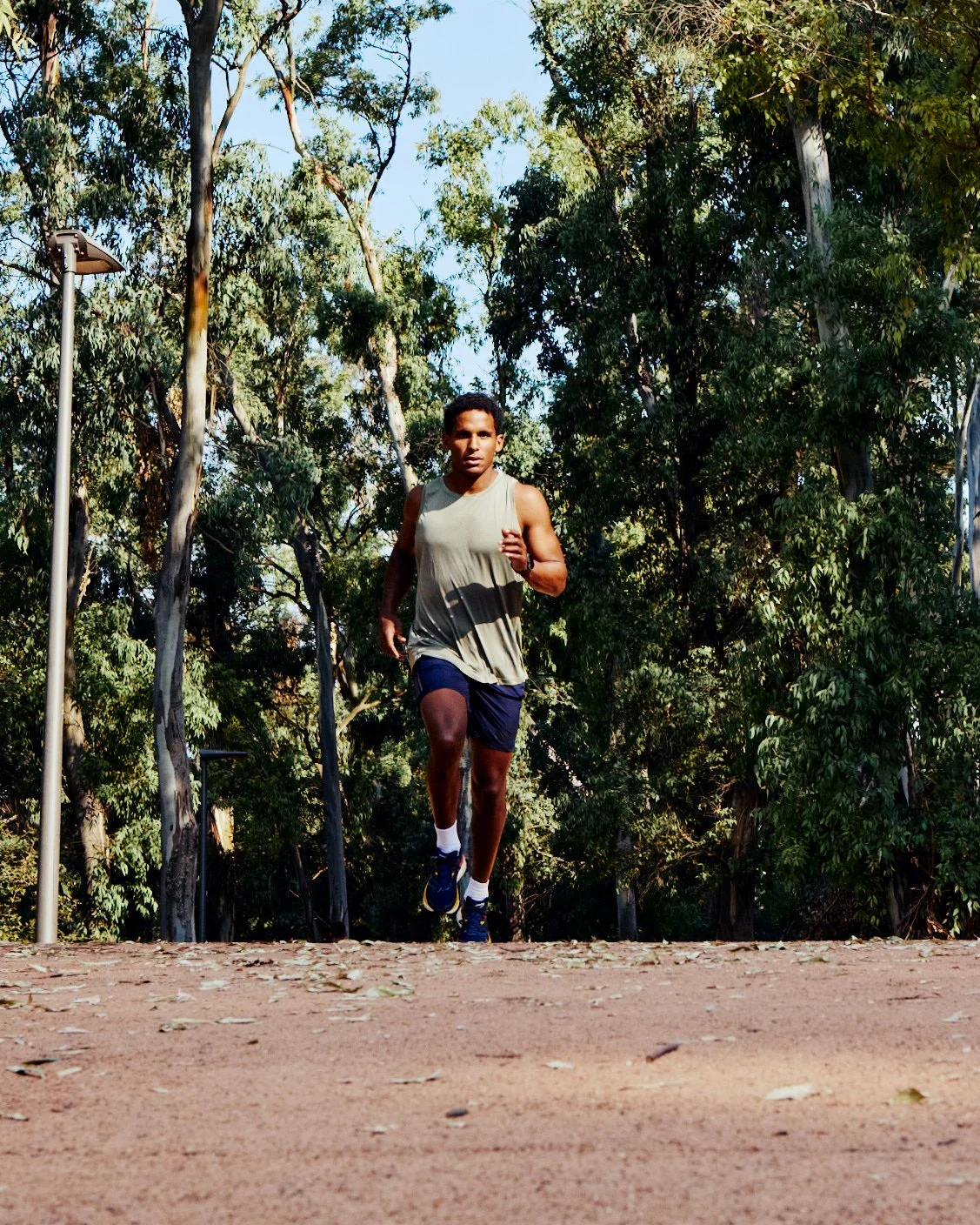 A young man running on an outdoor dirt trail surrounded by green trees and clear blue sky.