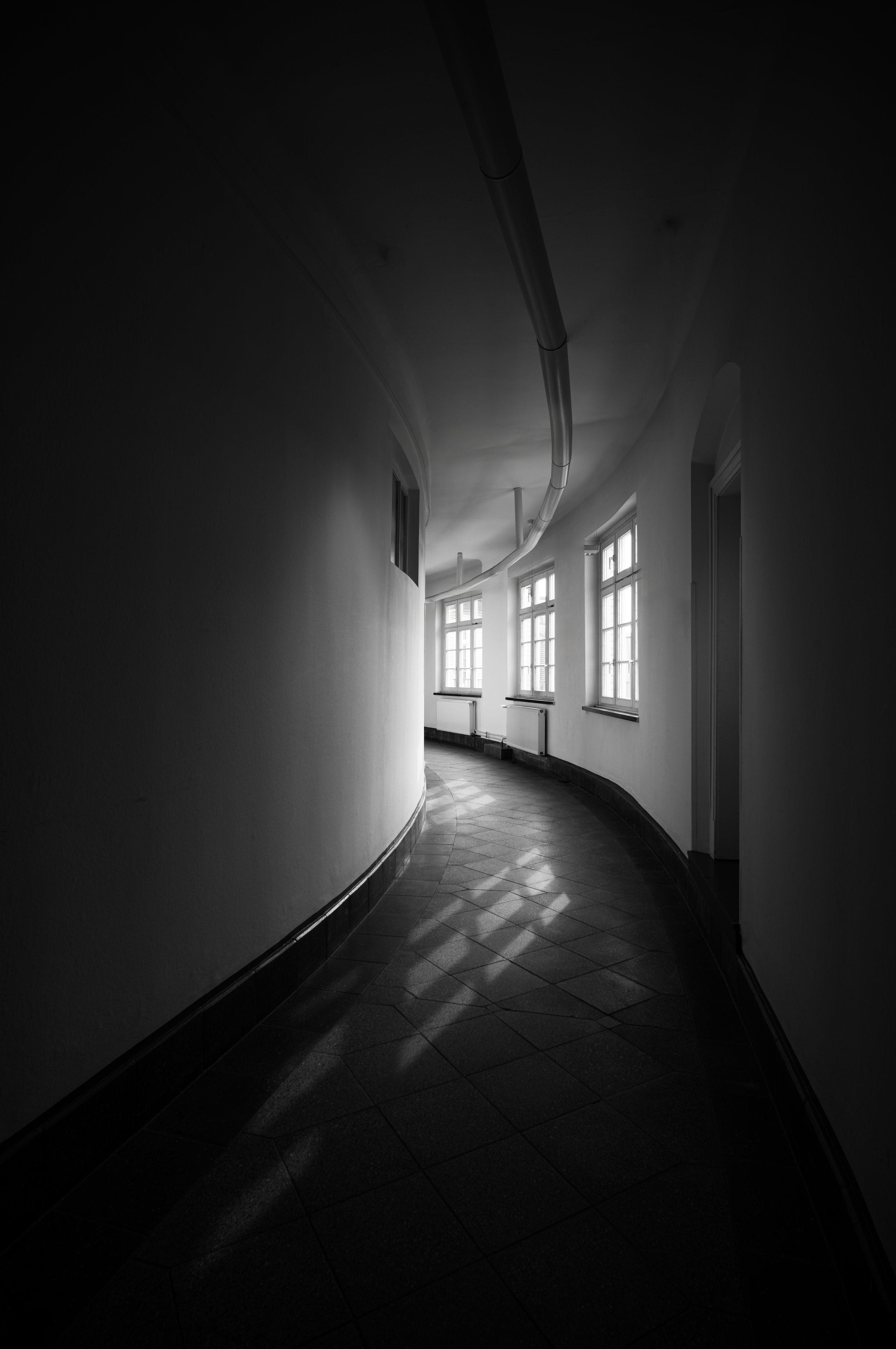 Ein dunkler, kurviger Flur mit mehreren großen Fenstern, die Tageslicht hineinlassen, und einem sichtbaren Rohr an der Decke.