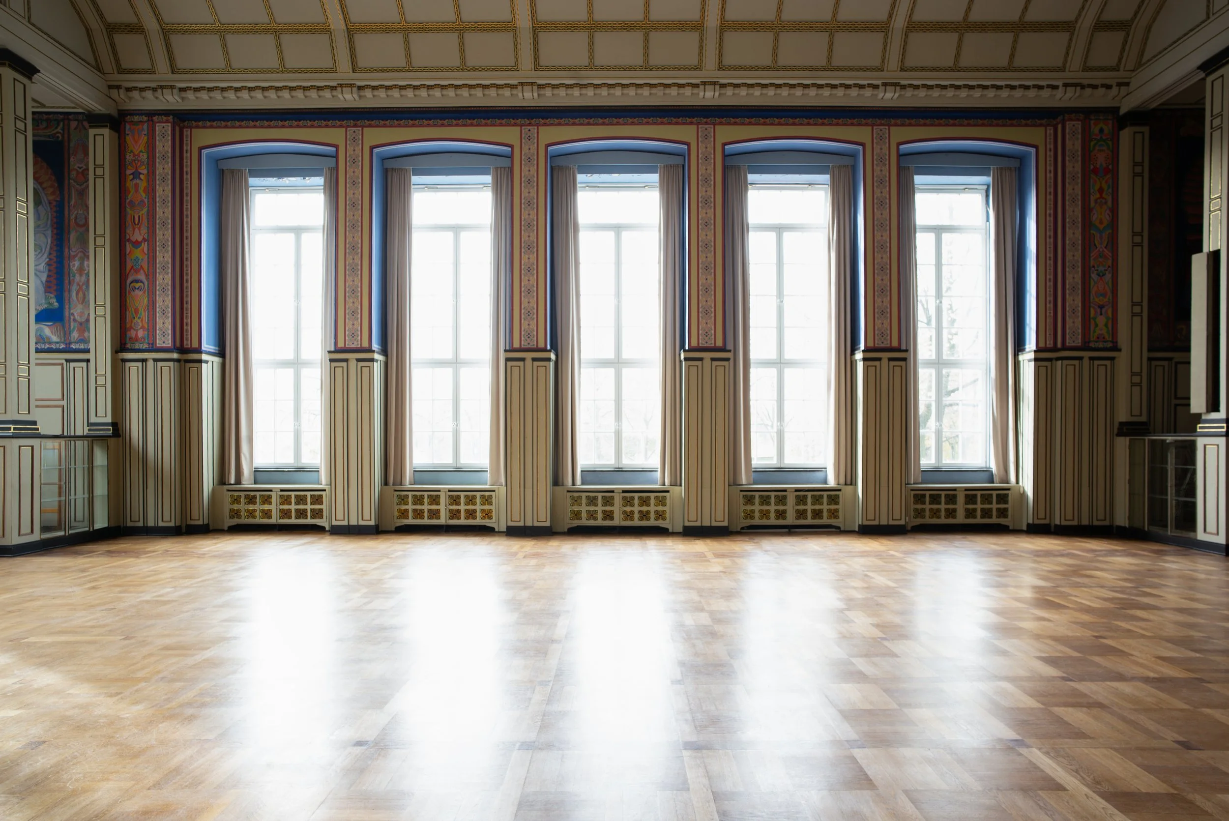 Ein großer, leerer Saal mit Holzparkett, hohen Fenstern und bunten Wandmalereien.