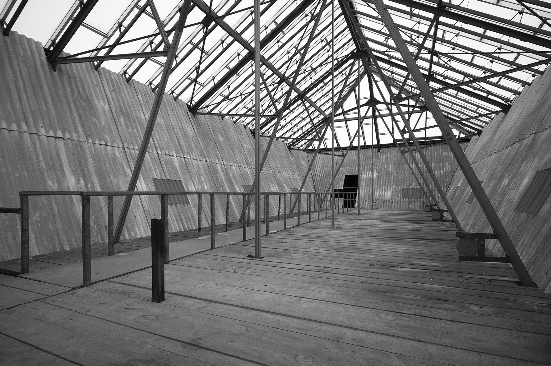 Ein leerer, alterndes Dachgeschoss mit einer Dachkonstruktion aus Metall- und Holzstrukturen, Staub und verrosteten Wänden, keine Möbel, das Licht kommt durch das Dach. Schwarz-Weiß-Bild.