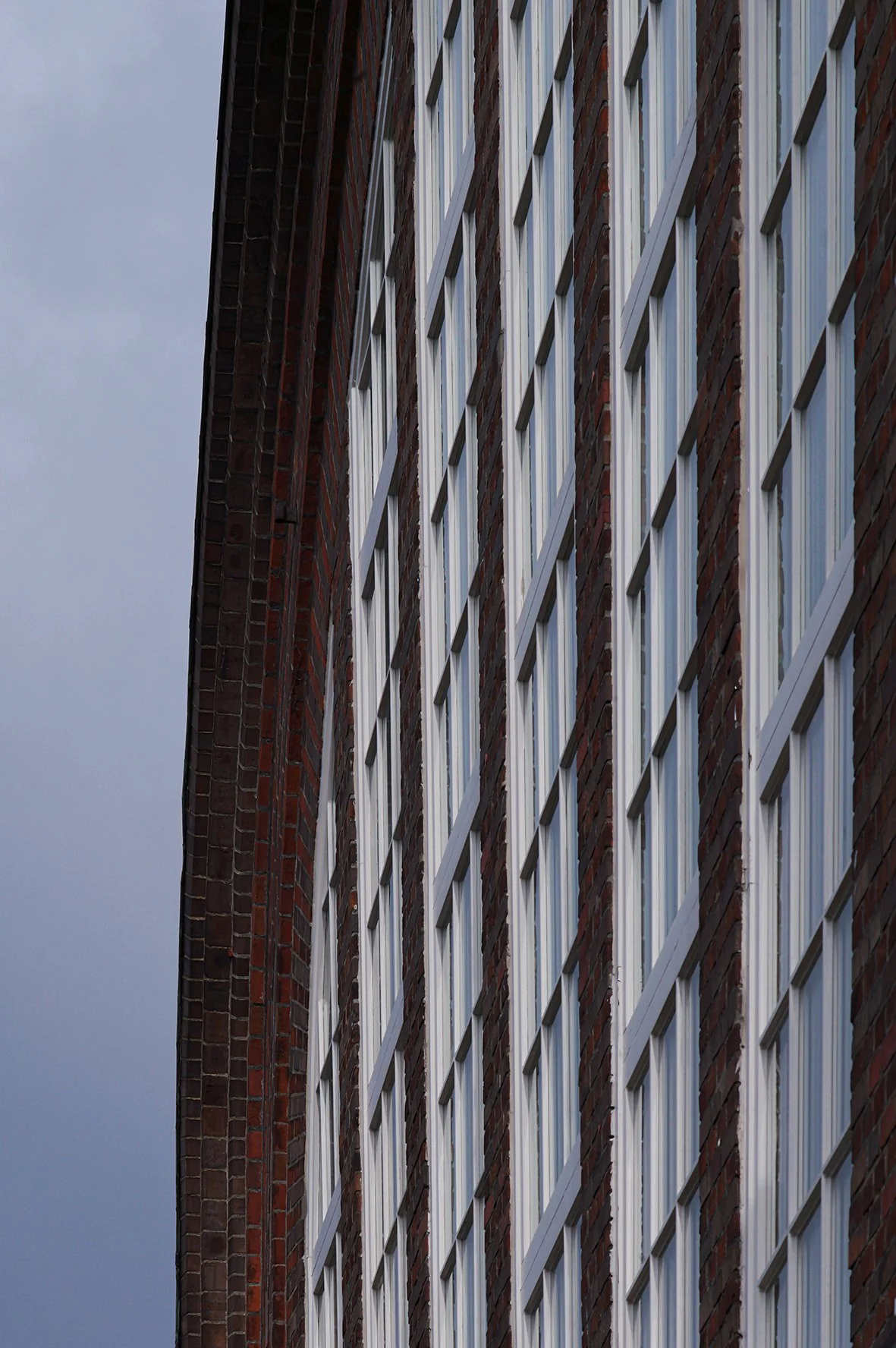 Außenansicht eines modernen Gebäudes mit großen rechteckigen Fenstern und rotem Ziegelmauer. Der Himmel im Hintergrund ist bewölkt.