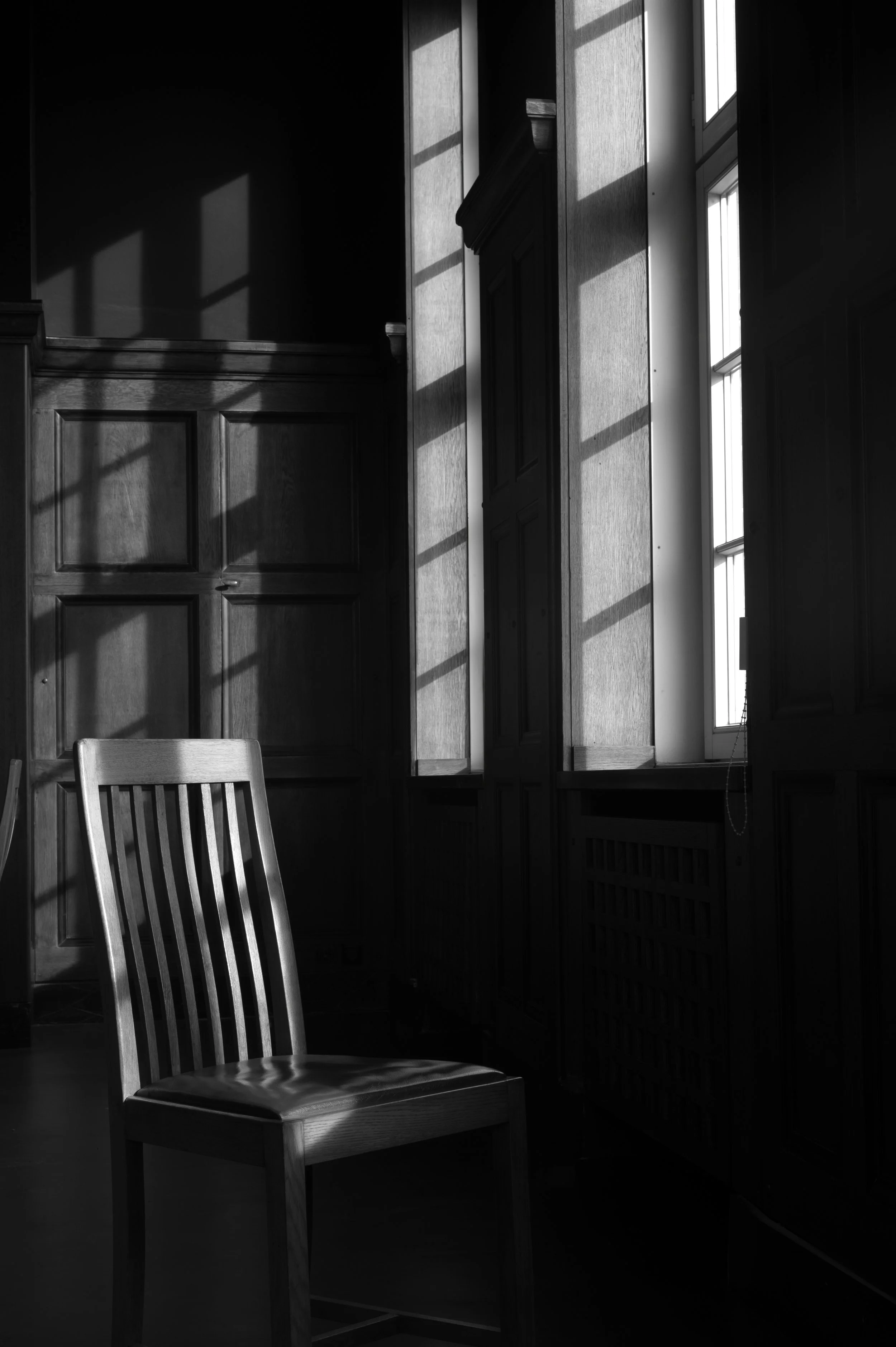Ein Holzstuhl in einem Raum mit großen Fenstern, durch die Sonnenlicht hereinfällt und Schatten wirft.