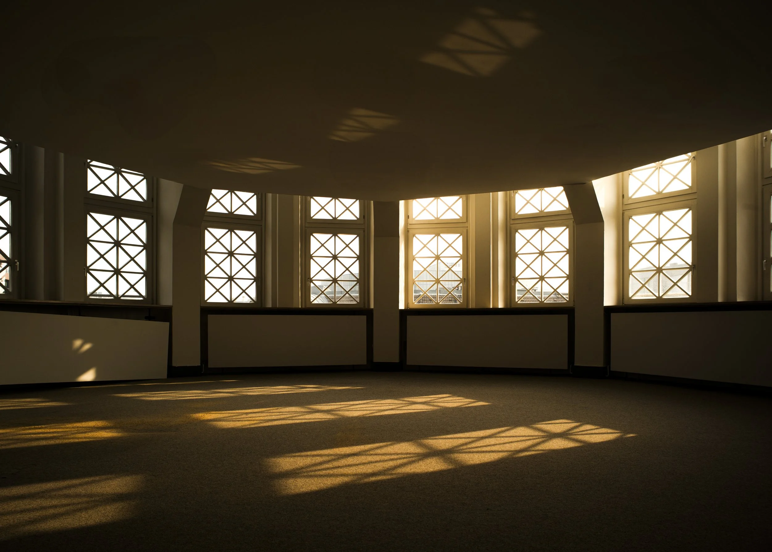 Ein leeres, lichtdurchflutetes Zimmer mit großen Fenstern, durch die Sonnenlicht und Schattenmuster auf den Boden fallen.