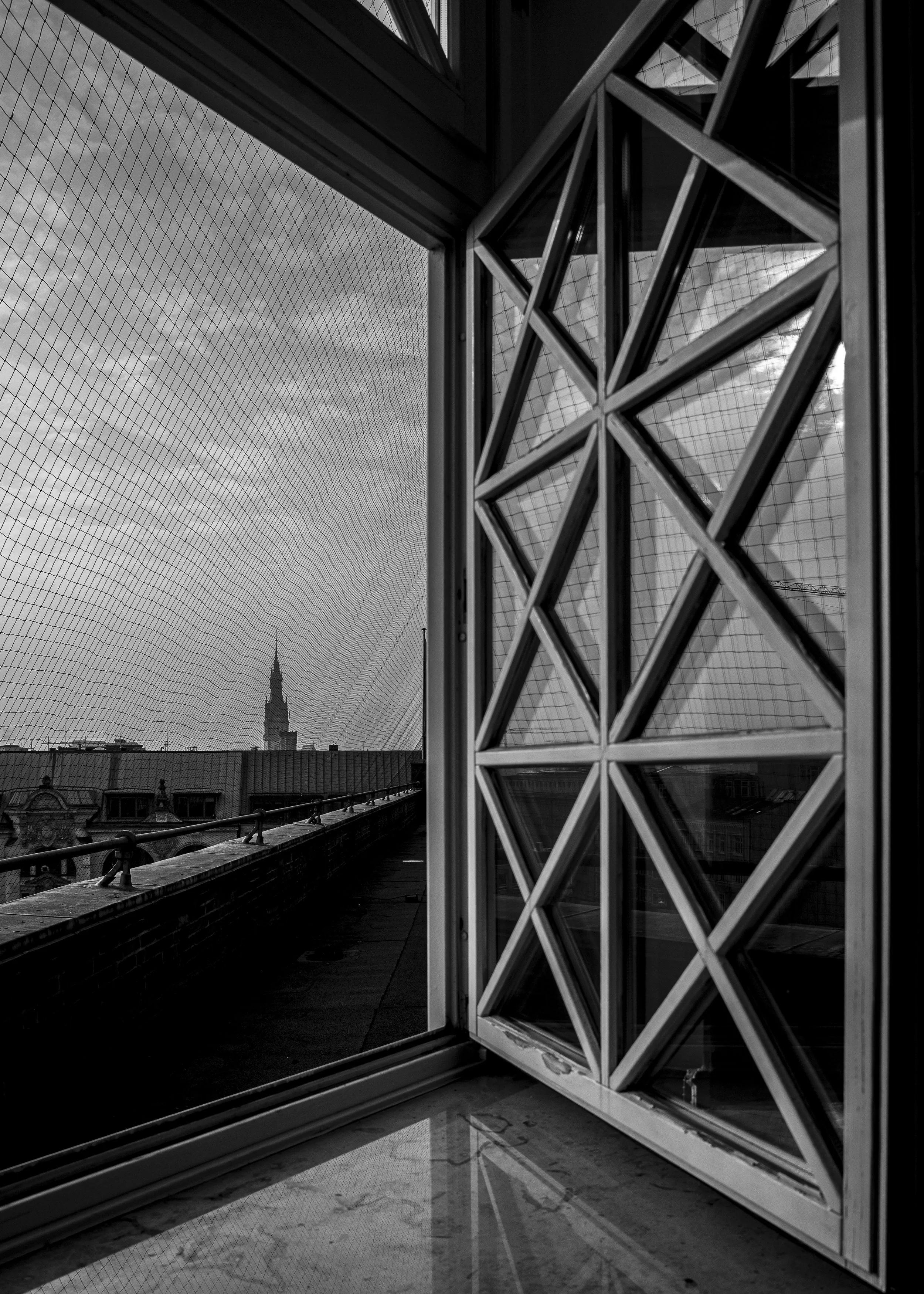 Schwarz-weiß-Foto von einem offenen Fenster mit einem Gitter und einem Blick auf eine Stadt mit Kirchturm im Hintergrund, bewölkter Himmel.