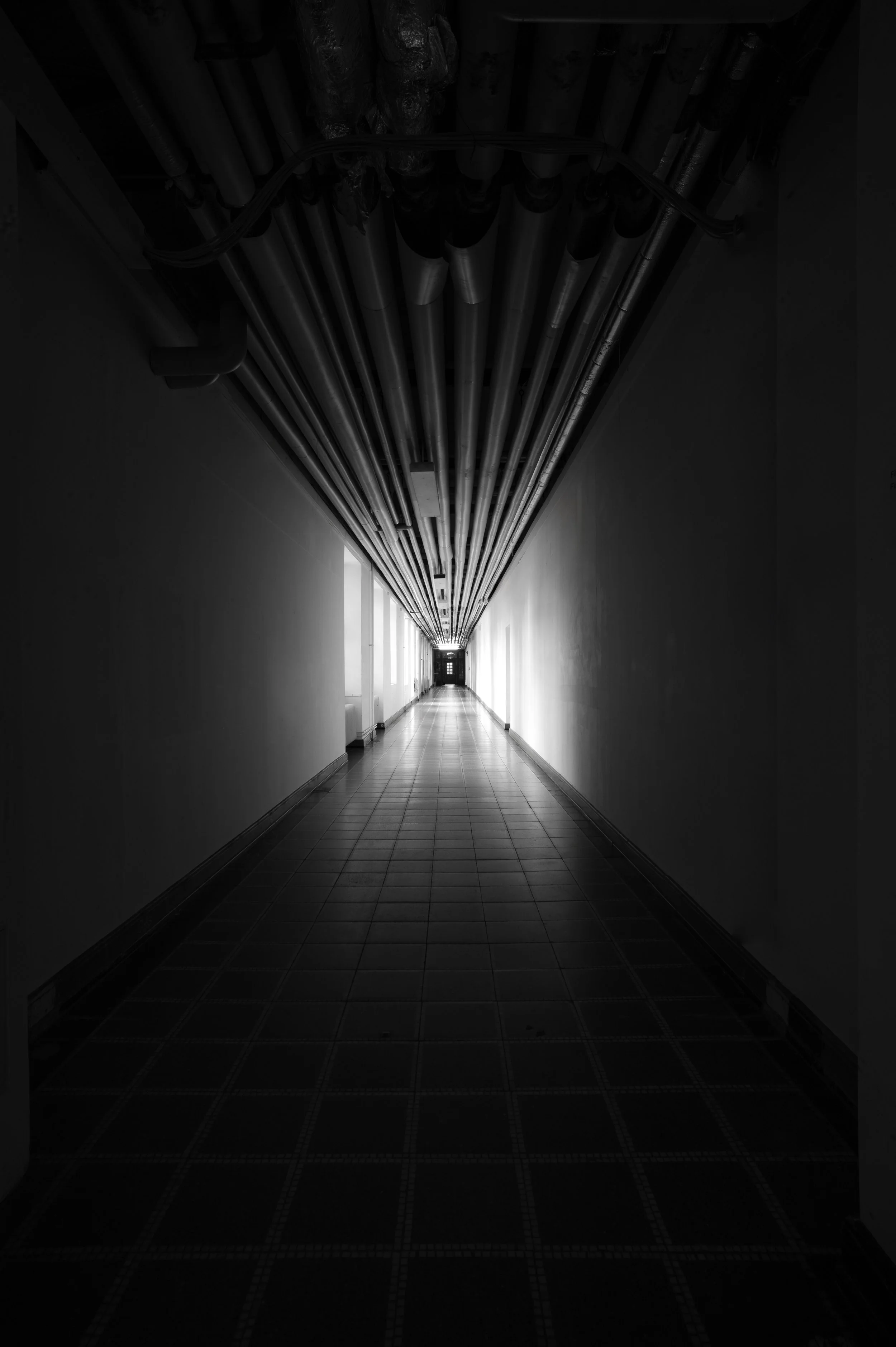 Ein langer, dunkler Flur mit weißen Wänden und einem Fliesboden, Deckenrohre sichtbar an der Decke.