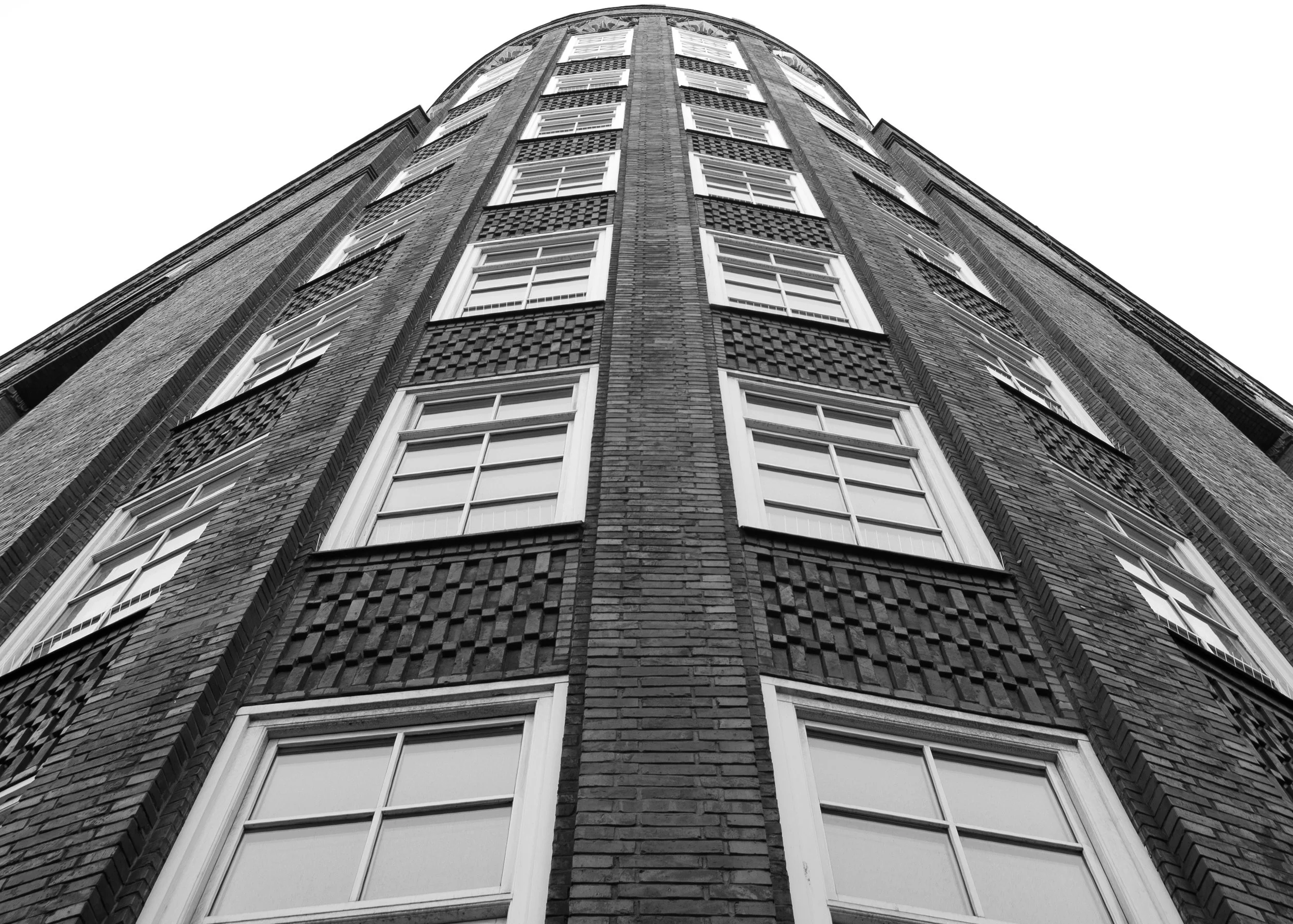 Aufnahme eines hohen, eher runden Gebäudes aus Ziegelsteinen im schwarz-weiß-Format, Blick von unten nach oben, mit mehreren Fenstern und schwarzen Gitterpartien an der Fassade.