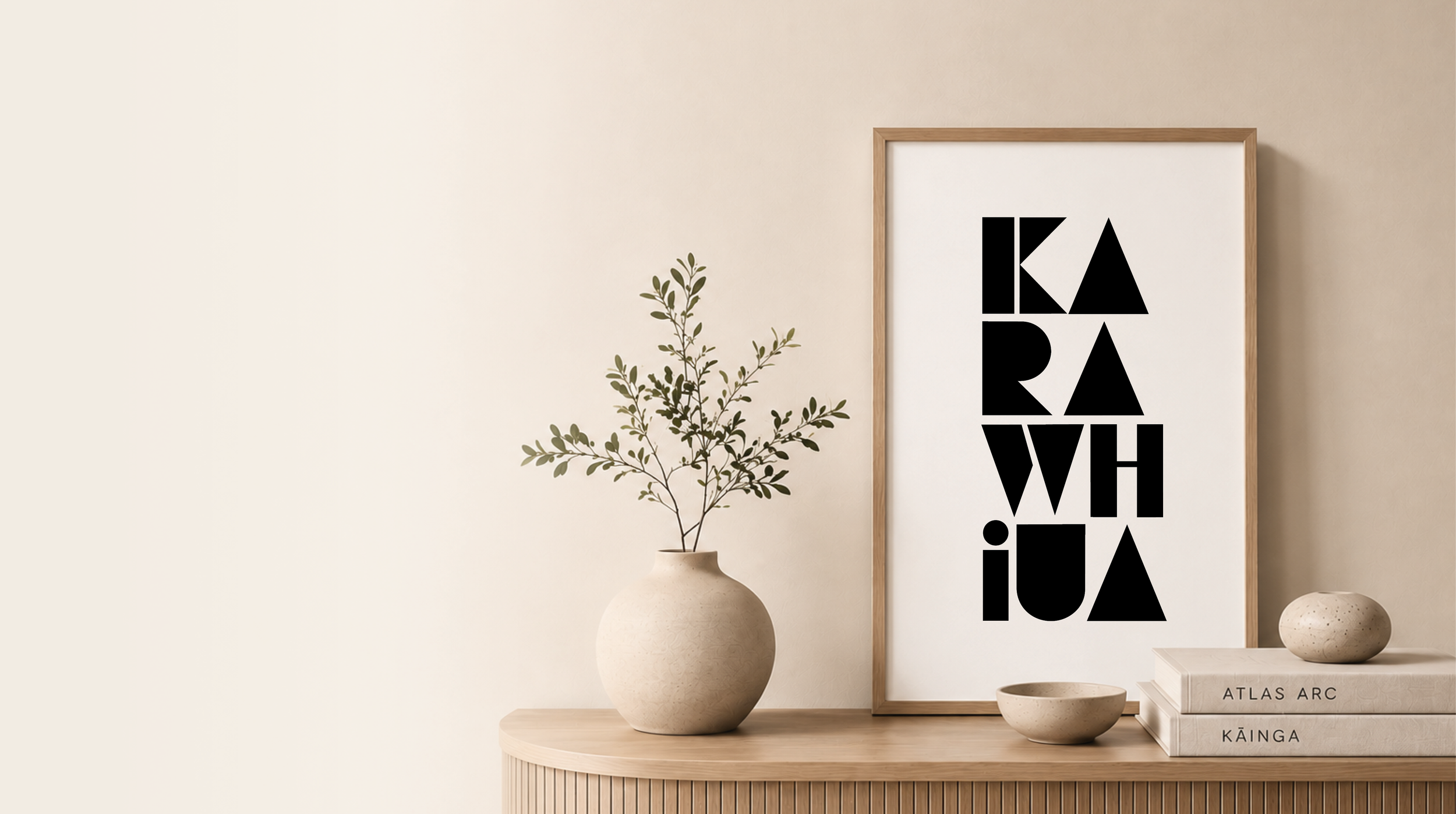 Decorative interior with a beige wall, a potted branch with green leaves, a framed art print with black geometric letters, two stacked books titled 'Atlas Arc' and 'Kainga', and small decorative bowls on a light wooden surface.