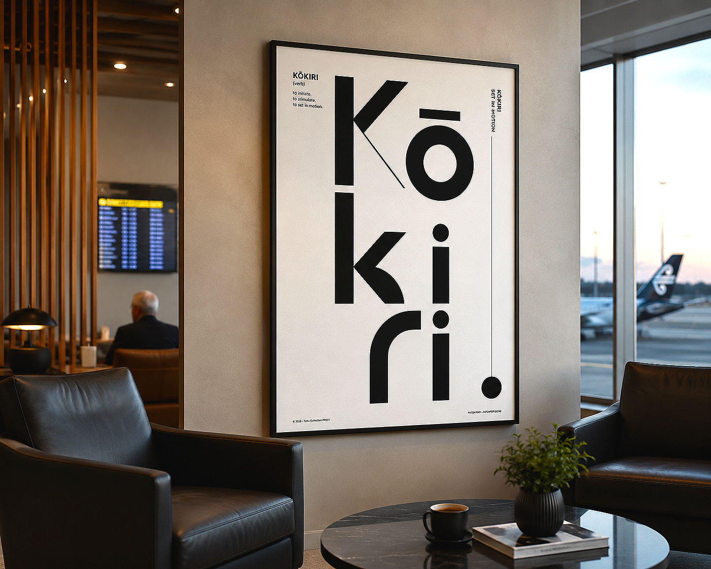 Interior of an airport lounge with a large black and white poster on the wall that says 'Kōkiri' with the definition 'to initiate, to stimulate, to set in motion'. There are black leather chairs, a small table with a black coffee mug, books, and a small plant. In the background, through large windows, airplanes are visible on the tarmac during sunset.