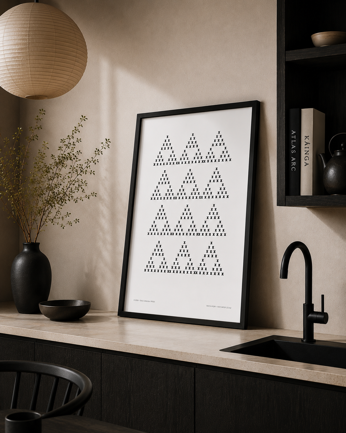Framed Māori pattern on a beige kitchen wall, with a black vase containing dried branches, a black bowl, black shelf with books, black faucet, and a black dining chair in the foreground.