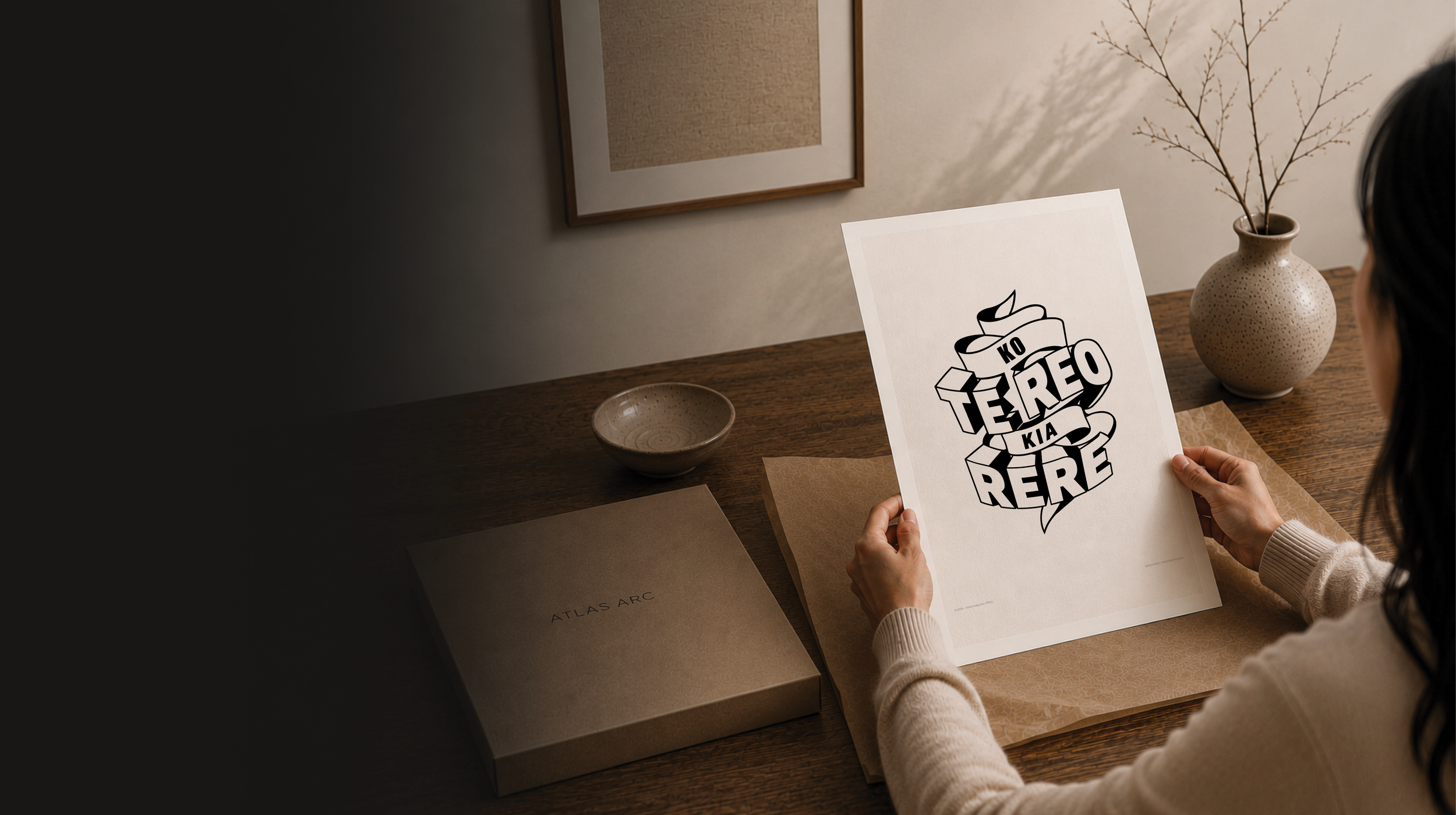 A person sitting at a wooden table holding a sheet of paper with black and white cartoon-style text that reads 'KO TE REO KIA RERE'. The table has a beige placemat, a ceramic bowl, and a boxed item labeled 'ATLAS ARC'.