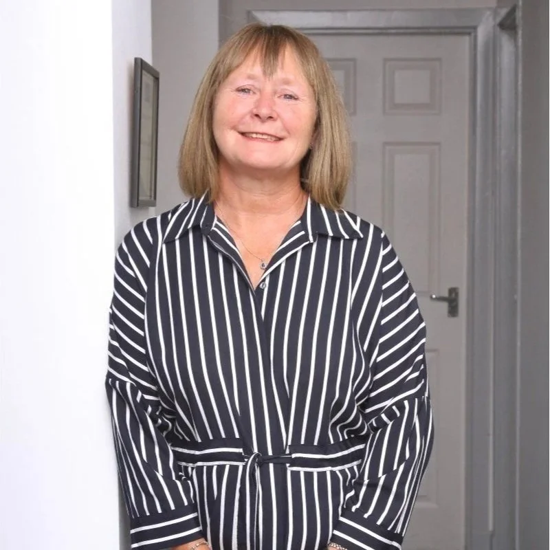 A smiling middle-aged woman with shoulder-length blonde hair wearing a black and white striped dress standing indoors against a light-colored wall and gray door.