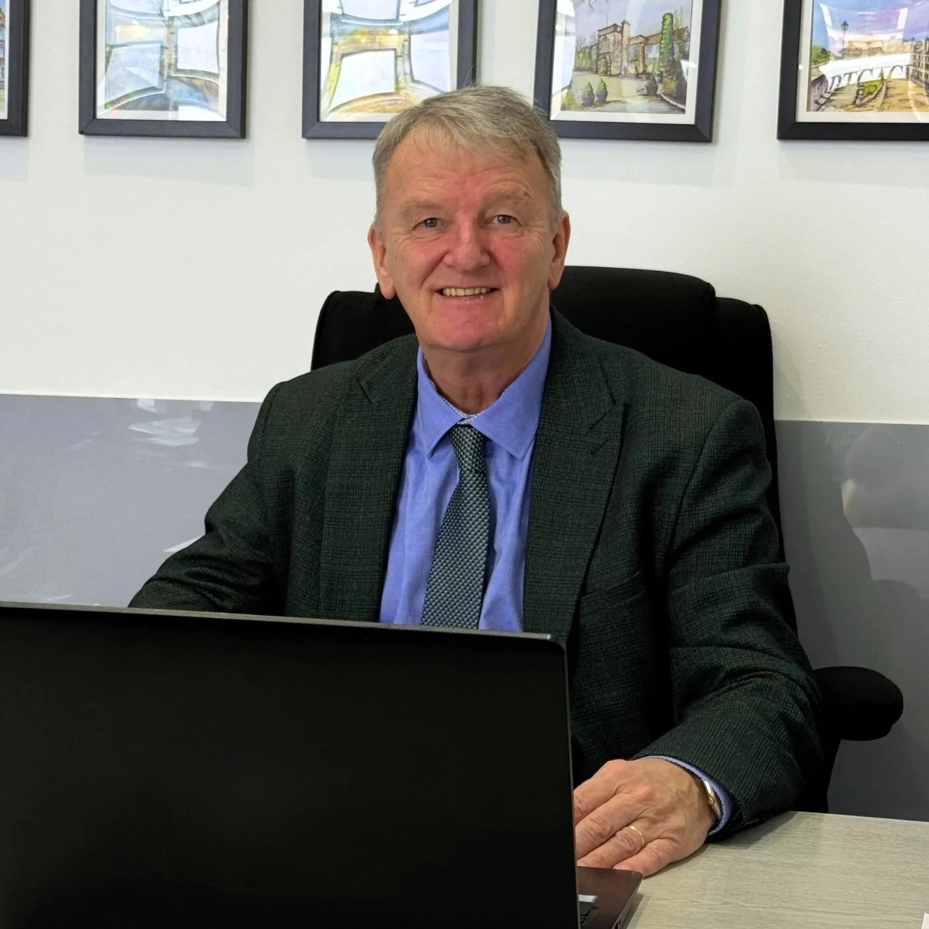 A smiling man in a business suit sitting at a desk in an office. Behind him are framed pictures of architectural scenes.