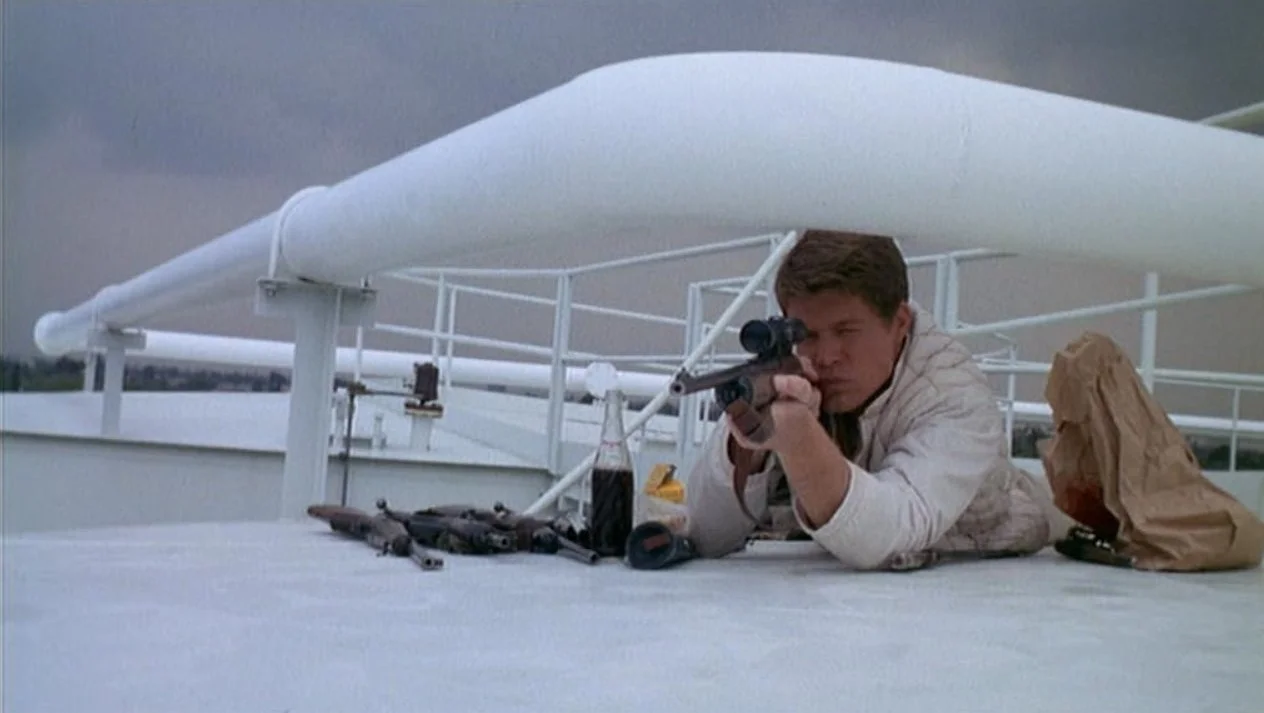 Man lying prone on a deck aiming a sniper rifle through a scope with a cloudy sky in the background. This is a scene from Peter Bogdanovich's debut 1968 film Targets, starring Boris Karloff.