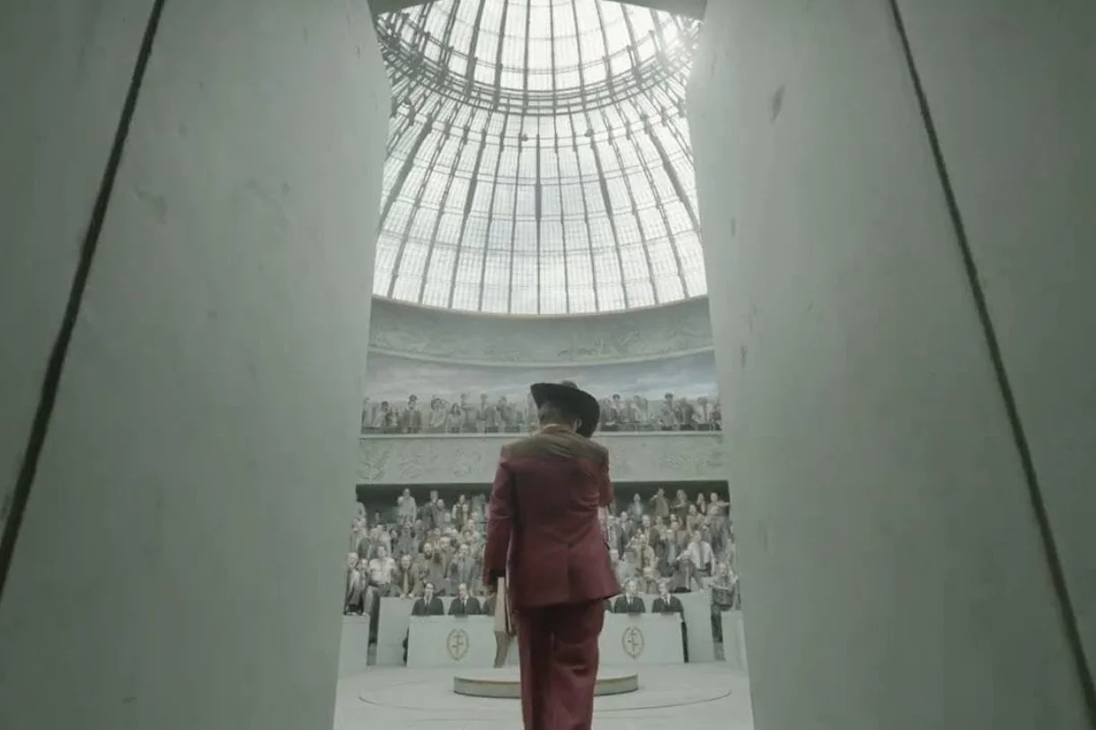 A man in a maroon suit stands in a large, domed assembly hall filled with seated people. The photo is taken from a low angle, with two pillars in the foreground framing the view upward to a glass ceiling. Digger (2026) Tom Cruise.
