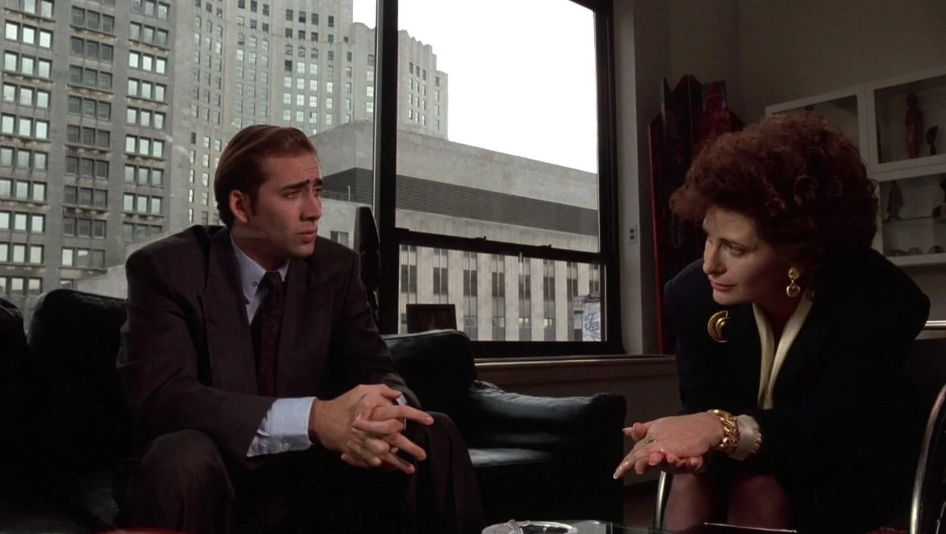 A man in a suit and a woman with curly hair and gold jewelry engaged in conversation in an office with large windows showing tall buildings outside. This is a still from the 1988 film Vampire's Kiss starring Nicolas Cage