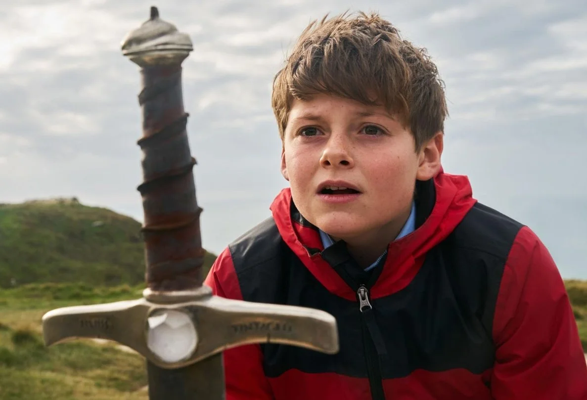 A film still from the family film The Kid Who Would Be King (2019) featuring Louis Ashbourne Serkis. A young boy wearing a black and red coat looking at an fancy sword.
