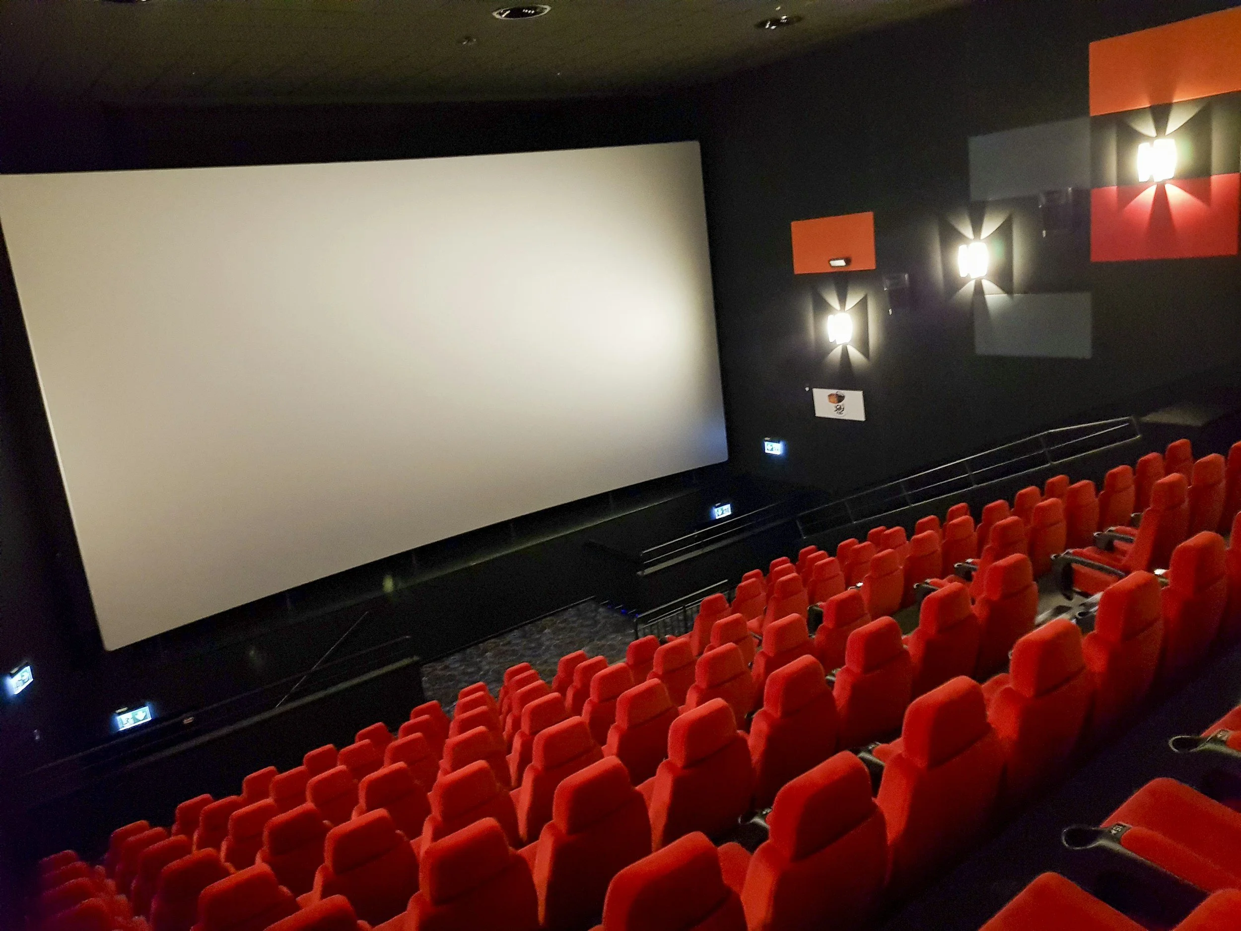 A movie theater with rows of red seats in front of a large blank white screen. The walls are dark, and there are wall-mounted lights with a decorative pattern. Exit signs are visible near the bottom of the seats.