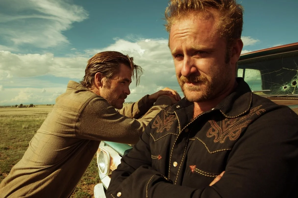 Two men posing by an old car with bullet holes on the window, set in an open field under a partly cloudy sky. This is a scene from the 2016 film Hell or High Water starring Chris Pine and Ben Foster.
