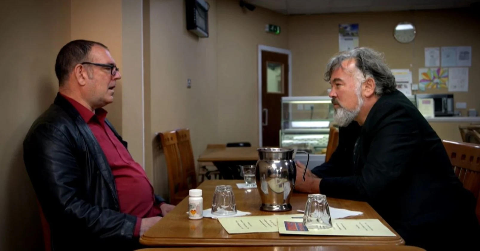Two men sitting across from each other at a dining table having a conversation in a restaurant or cafe. One man has gray hair and a beard, the other man wears glasses and has short dark hair. This is a scene from the 2020 Documentary film King Rocker