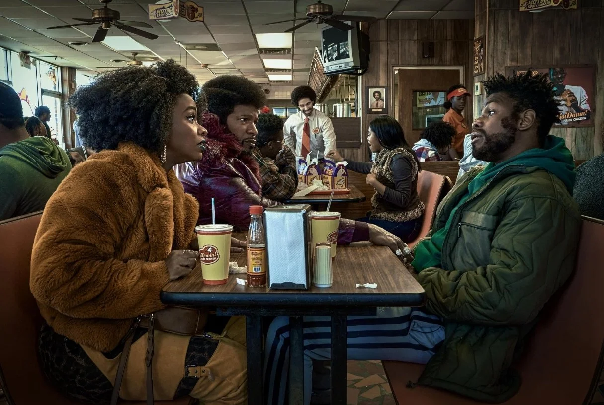 A group of people sitting at a table in a casual restaurant, engaging in a serious conversation. The background shows other patrons and a server.
They Cloned Tyrone (2023) film directed by Juel Taylor, Starring Jamie Foxx and John Boyega.