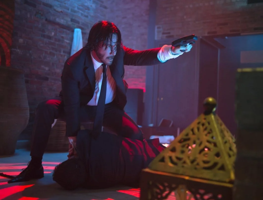 A man in a suit holding a gun and in a crouched attack position over another person lying on the floor, in a dimly lit room with red and blue lighting. This is a still from the 2014 film John Wick directed by Chad Stahelski and starring Keanu Reeves.