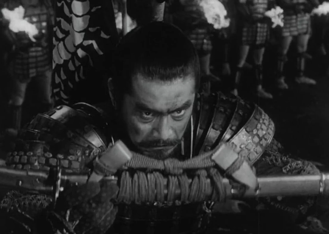 Black and white image of a man in traditional Japanese samurai armor, looking intensely forward while holding a samurai sword. This is a still from the Akira Kurosawa film Throne of Blood (1957) starring Toshiro Mifune