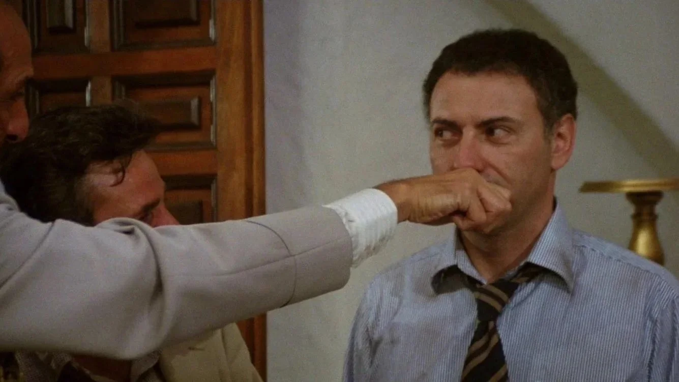 A film still from the comedy adventure film The In-Laws Featuring Alan Arkin, he is being shown a mans hand with a face drawn on it (this is not shown) Arkin is trying not to laugh..