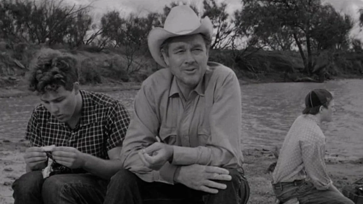 A film still from The Last Picture Show (1971) starring Timothy Bottoms, Ben Johnson and Sam Bottoms. Three men sitting by a riverbank, one with a cowboy hat talking.
