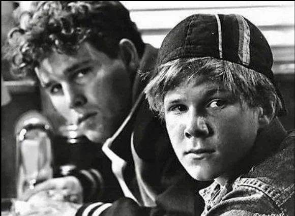 Black and white film still from 1971 film The Last Picture Show, starring Timothy Bottoms and Sam Bottoms, two teen boys sitting closely together, one wearing a baseball cap backwards.