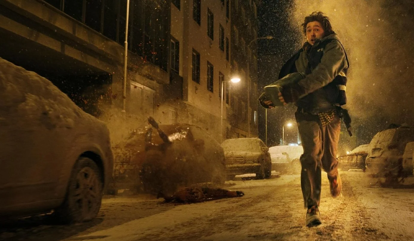 A person running on a snowy city street at night, holding a book or object, with cars covered in snow around, and buildings lit by streetlights. Resident Evil (2026) directed by Zach Cregger and starring Austin Abrams.