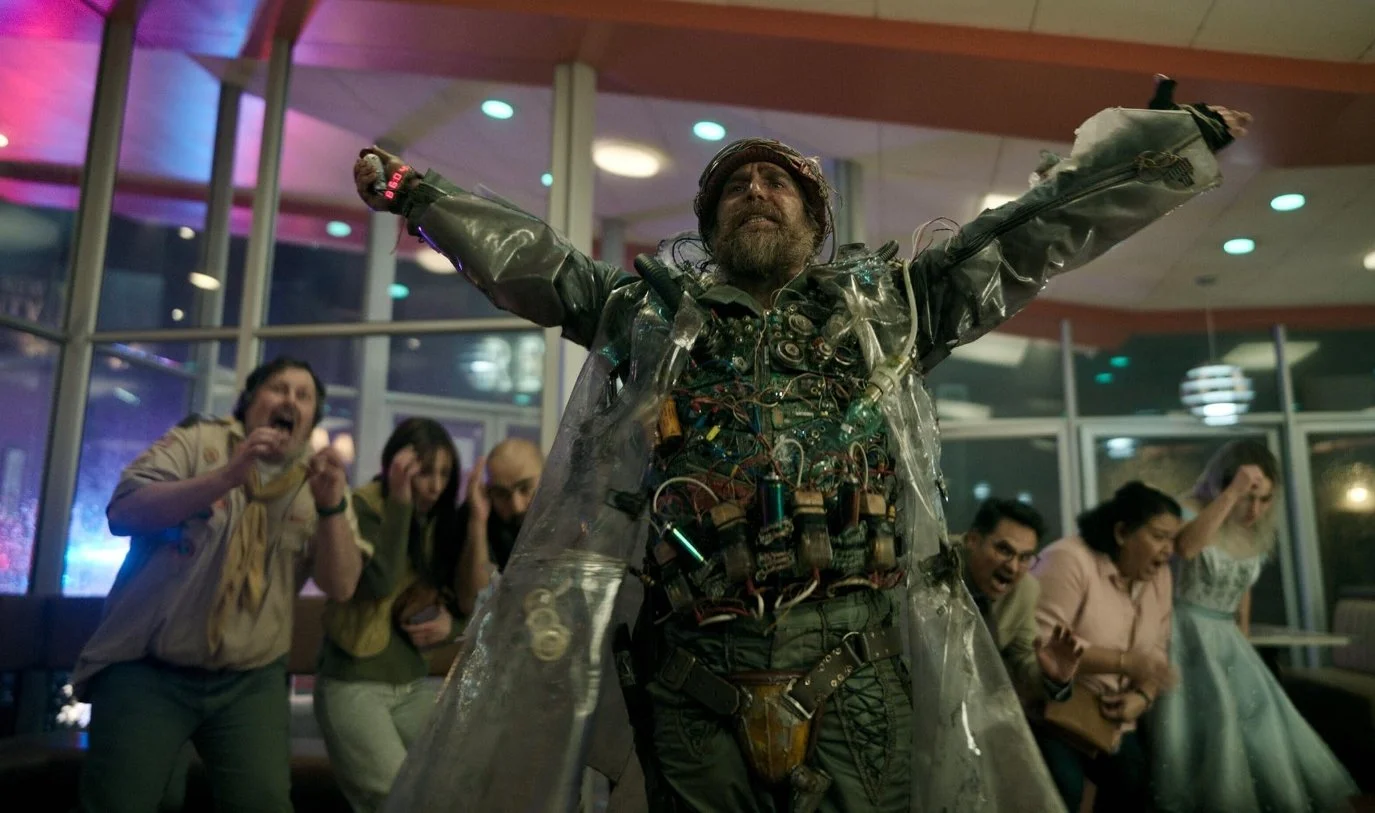 Bearded man in a makeshift wired costume raises his arms while frightened people recoil indoors. Good Luck, Have Fun, Don't Die (2026) directed by Gore Verbinski and starring Sam Rockwell, Juno Temple and Haley Lu Richardson.