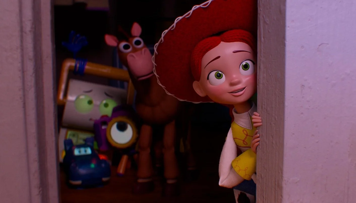 Animated cowgirl with red hair and green eyes peeking around a corner, with various toys in a dimly lit room behind her, including a horse, a cube with eyes, and a robot. From Toy Story 5 (2026), starring Joan Cusack, Tom Hanks, Greta Lee, Tim Allen.