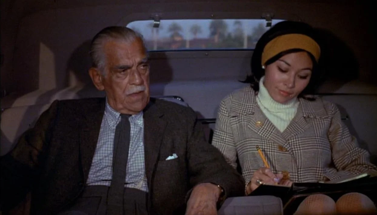 An older man and a young woman sitting in the back of a car. The man has white hair and is wearing a dark suit, while the woman has black hair with a yellow headband and is writing in a notepad. Targets (1968) Bogdanovich