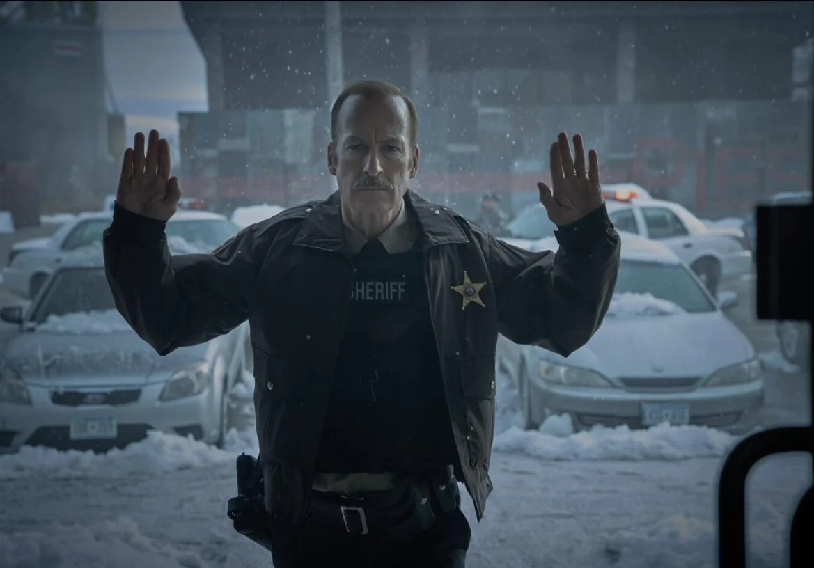 A sheriff's deputy stands outside on a snowy day, pressing his hands up in the air in front of a glass door, looking into a building. This is a still from the 2025 action film Normal directed by Ben Wheatley starring Bob Odenkirk.